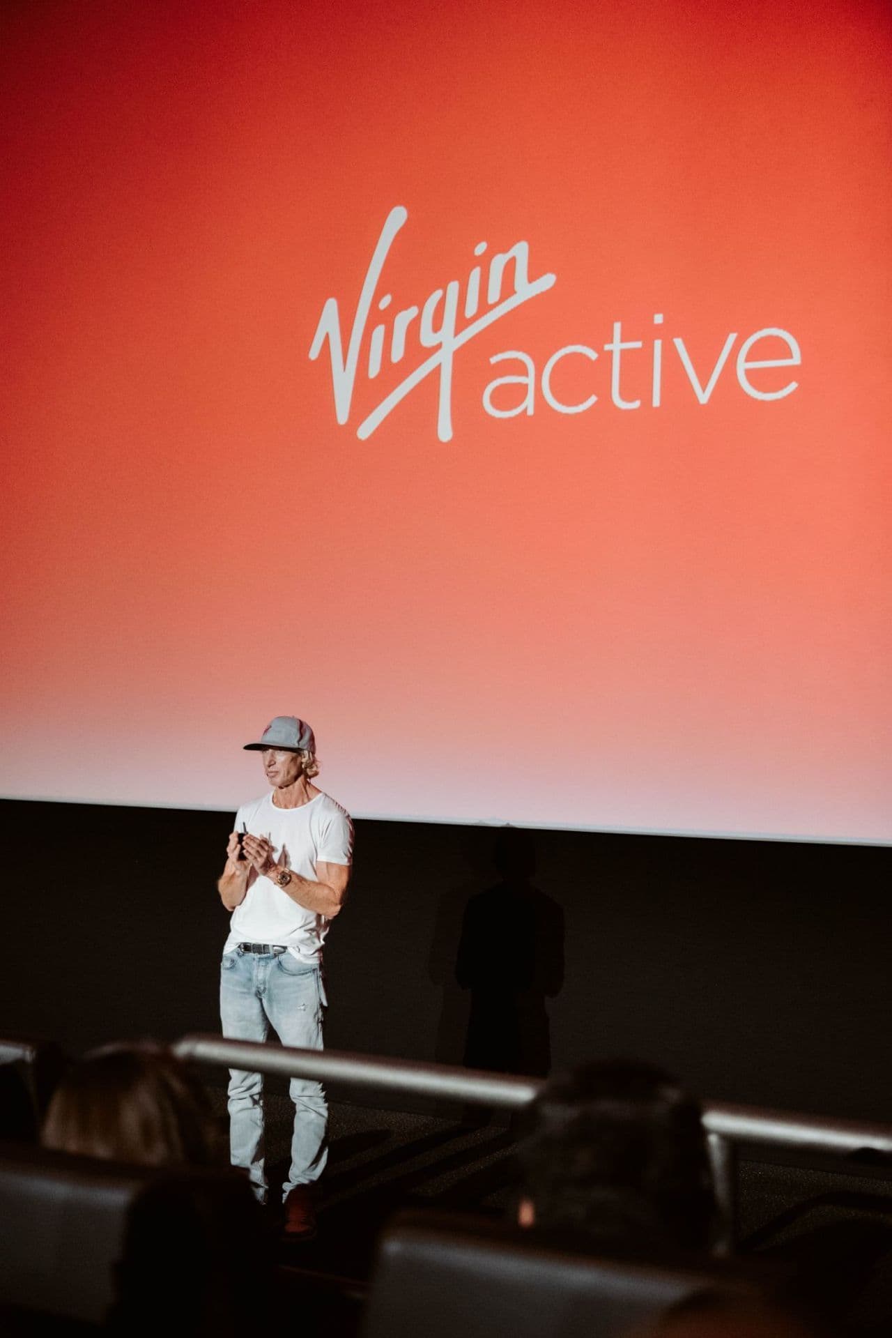 A male speaker presenting on stage in front of a large red Virgin Active screen to an audience.