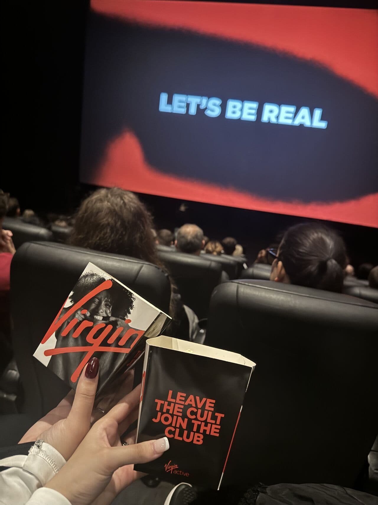 Person holding Virgin Active branded snack boxes in a cinema, with "Let's be real" displayed on the screen.