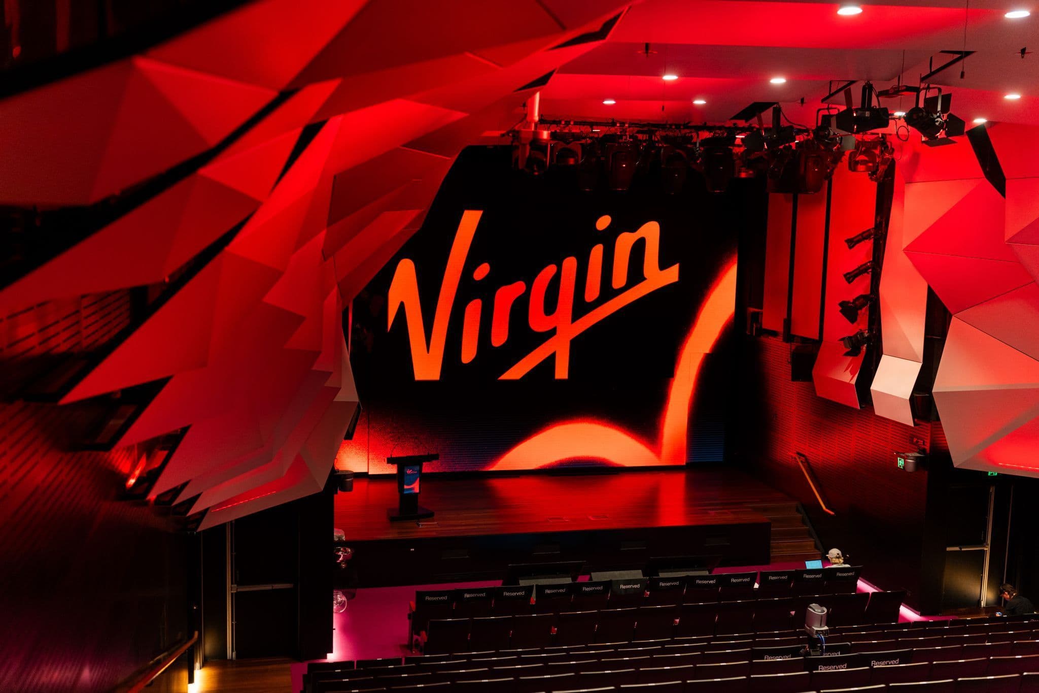 Modern auditorium with geometric wall panels, empty seating, and a stage featuring a large Virgin logo on the screen.