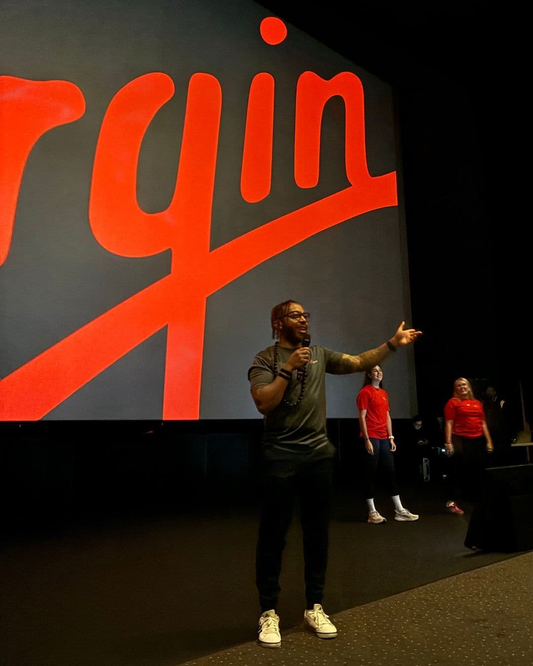 A male host speaking into a microphone on stage at a Virgin event, with a giant red Virgin logo in the background.