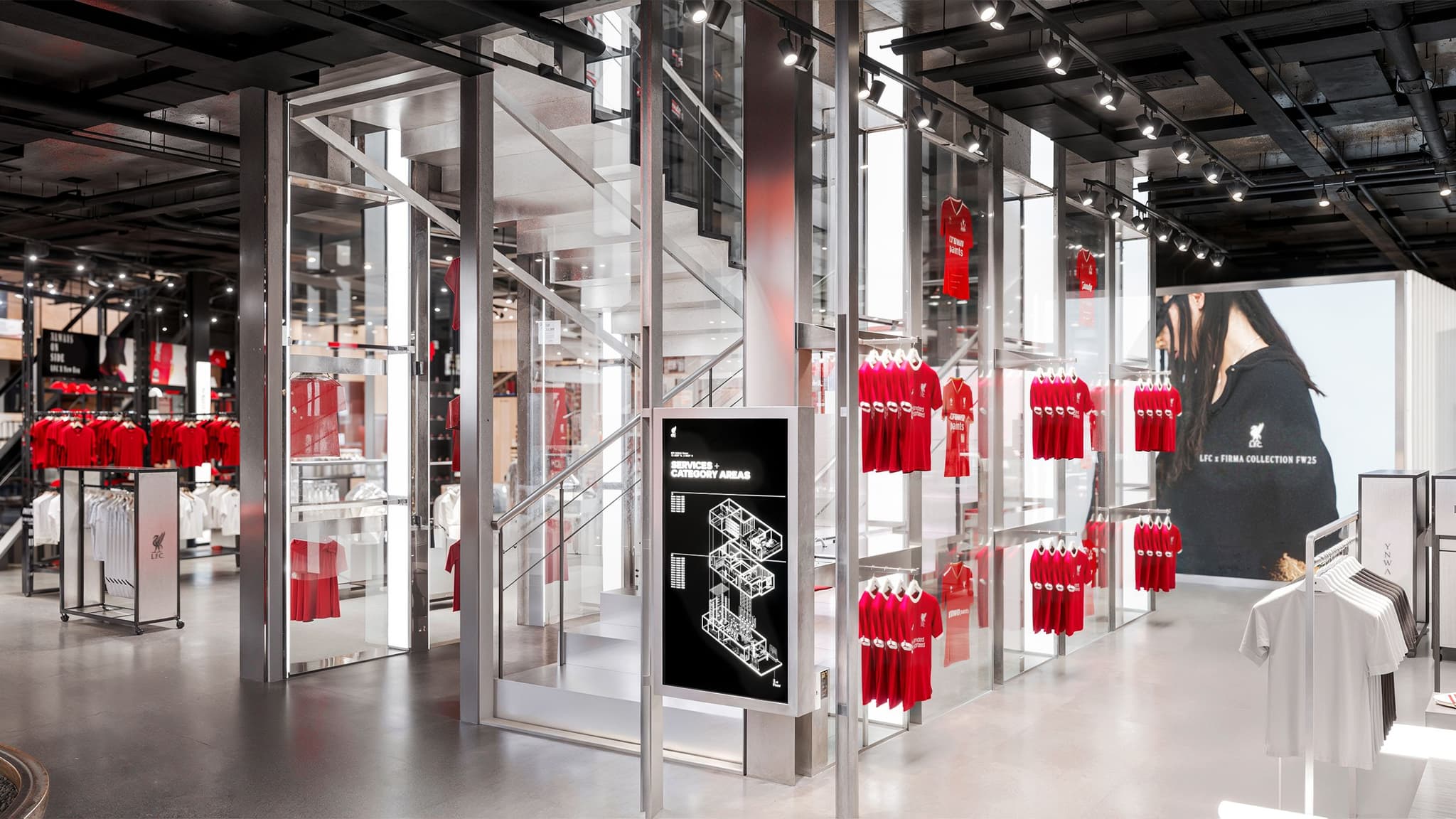 Modern glass staircase and digital store directory inside a Liverpool FC retail store with red shirts on display.