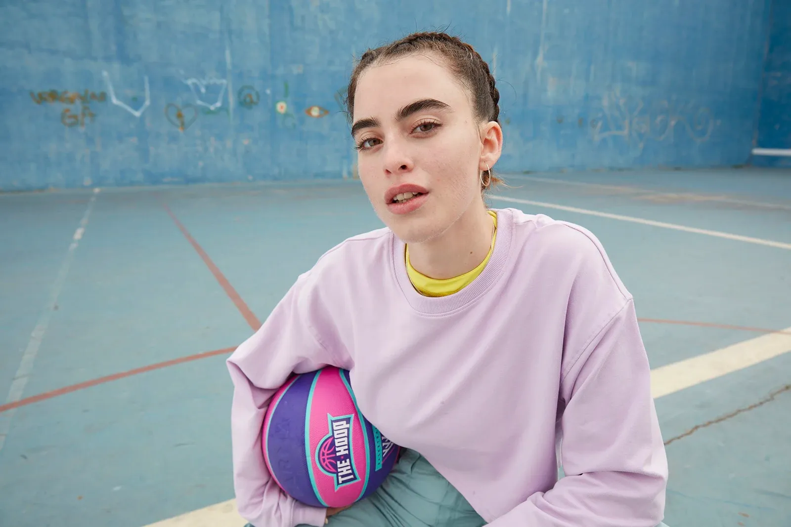 Young woman in a pink sweater sitting on a blue outdoor basketball court holding a colorful pink and purple basketball.