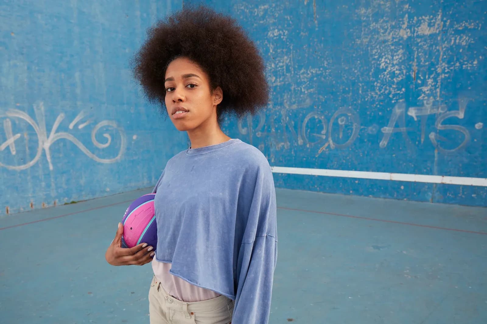 Young woman standing on a blue basketball court holding a colorful basketball against a blue wall.