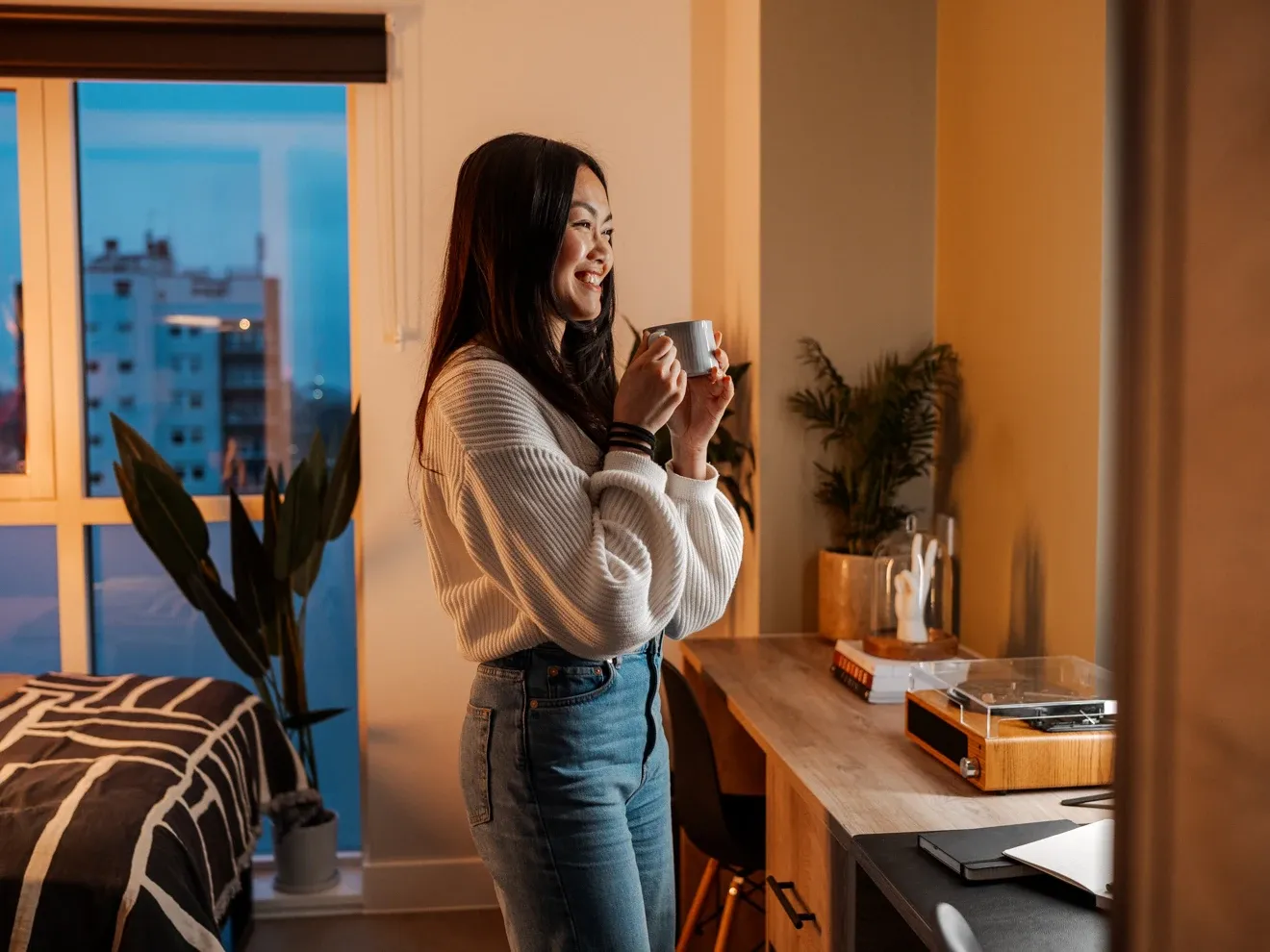 Smiling woman holding a coffee mug while standing by a desk in a modern student bedroom at dusk.