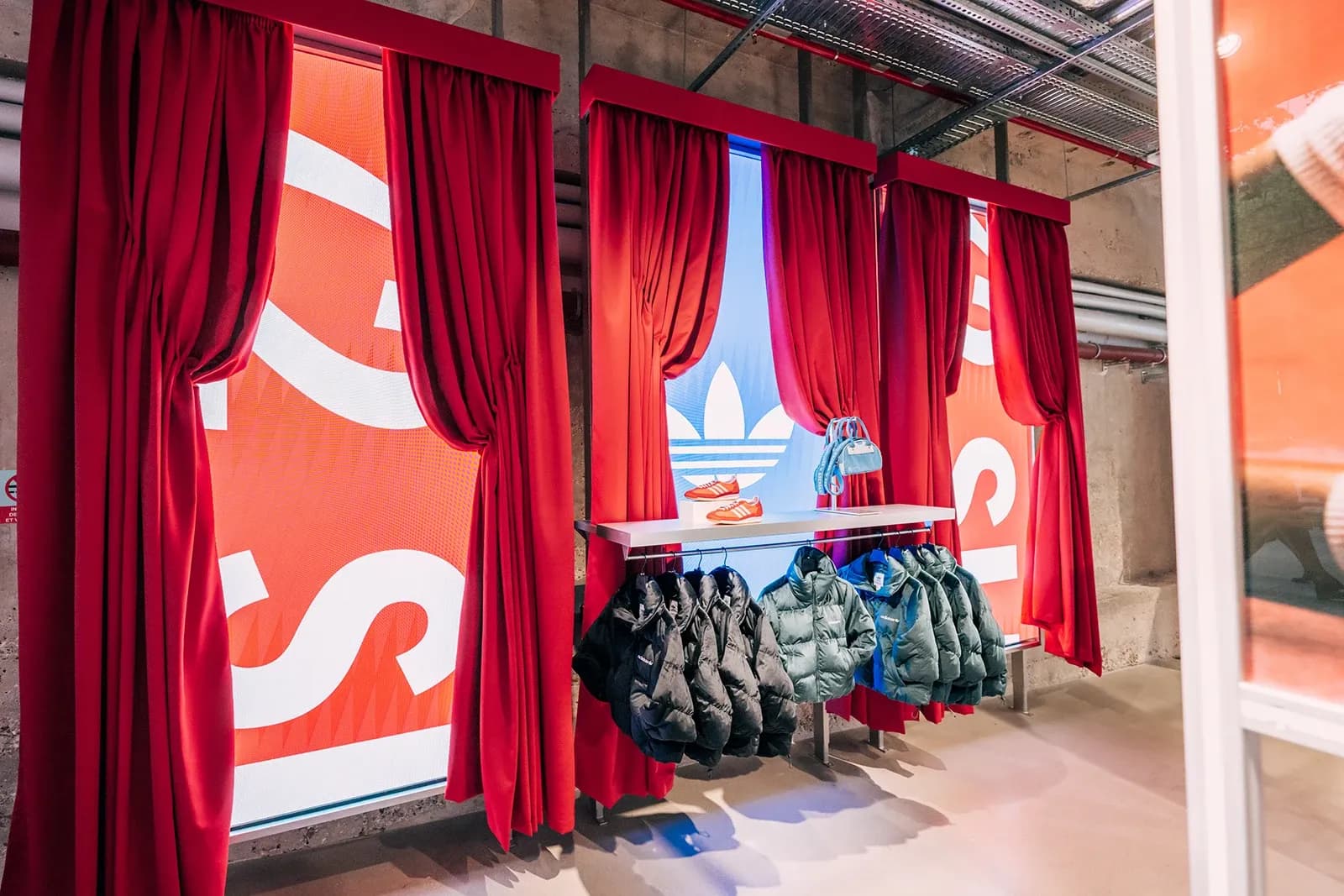 Clothing rack displaying green and black Adidas puffer jackets in front of red curtains and a trefoil logo screen.