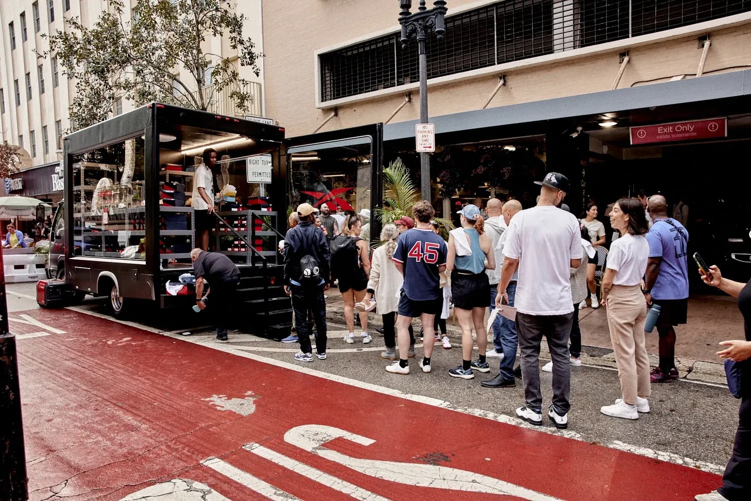 Crowd of people lining up on a city sidewalk next to a parked black Reebok glass-sided promotional truck.