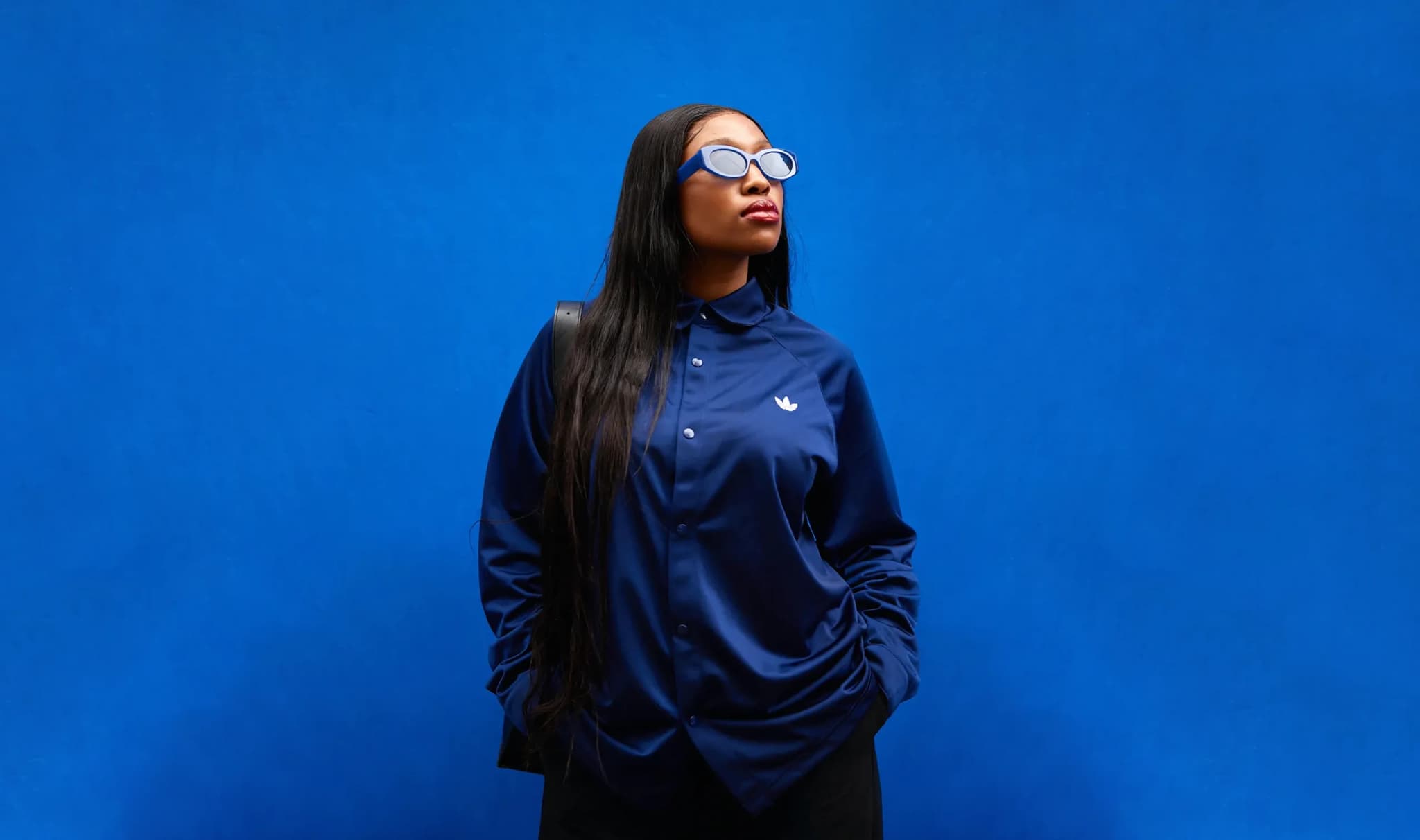 Person posing in a dark blue button-up shirt and sunglasses against a matching solid blue background.