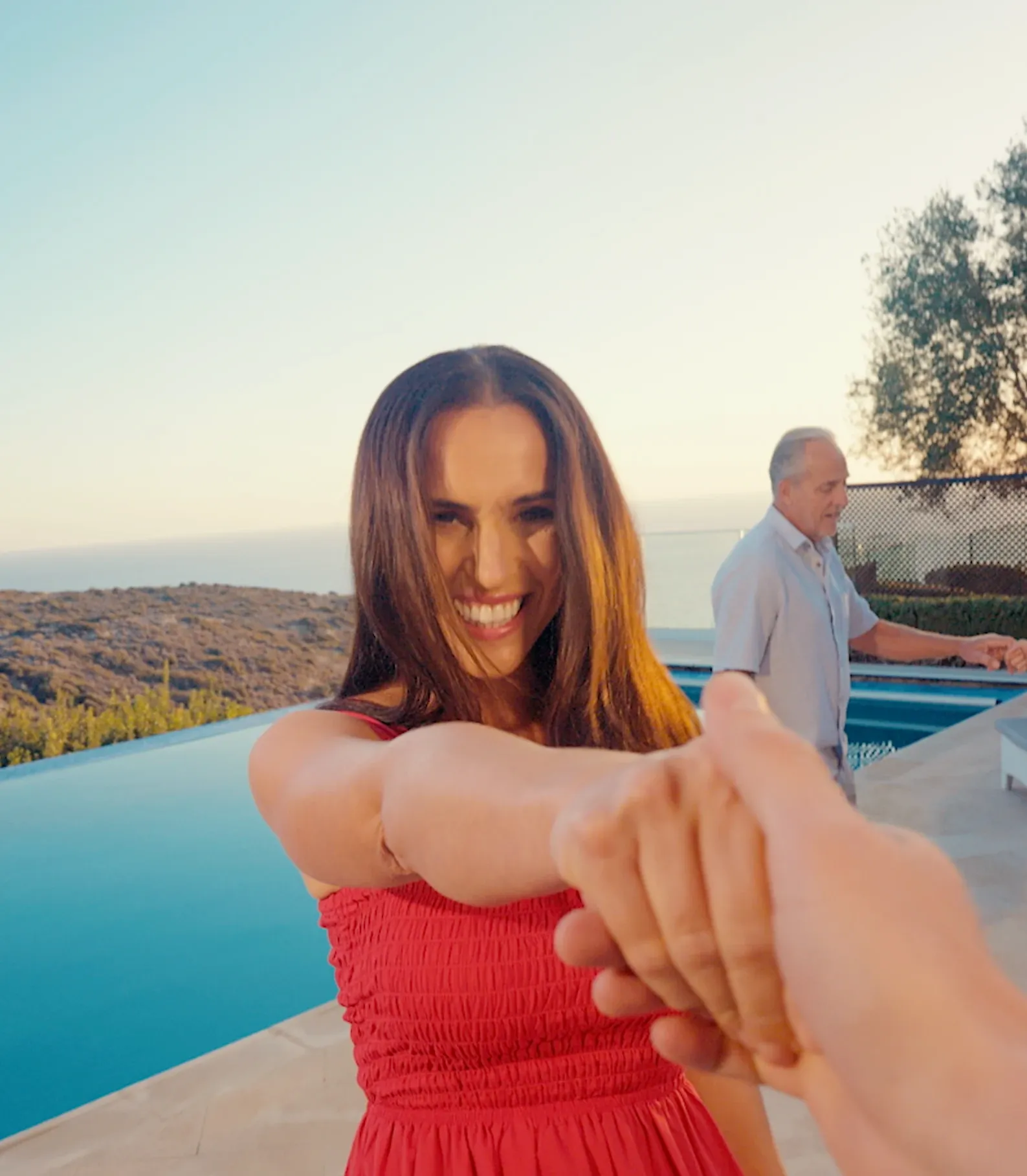 POV shot holding hands with a smiling woman in a red dress by a pool at sunset, with a man dancing in the background.