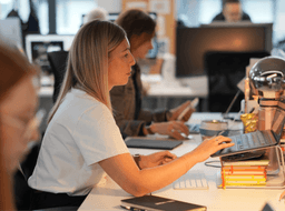 Two women sitting side-by-side working on laptops in a focused, collaborative office environment.