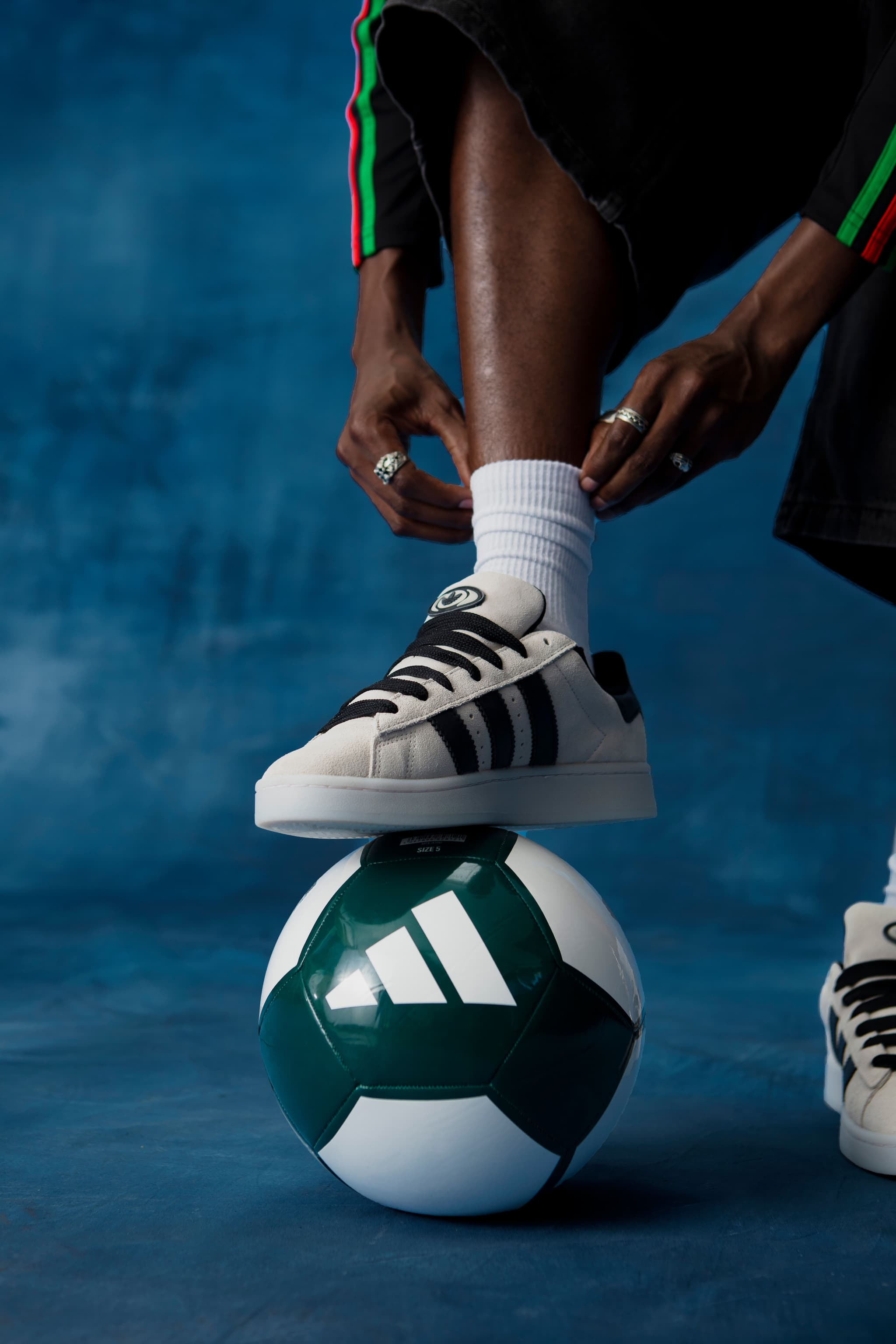 Close-up of a person tying a white and black adidas Campus sneaker resting on a green and white adidas soccer ball.