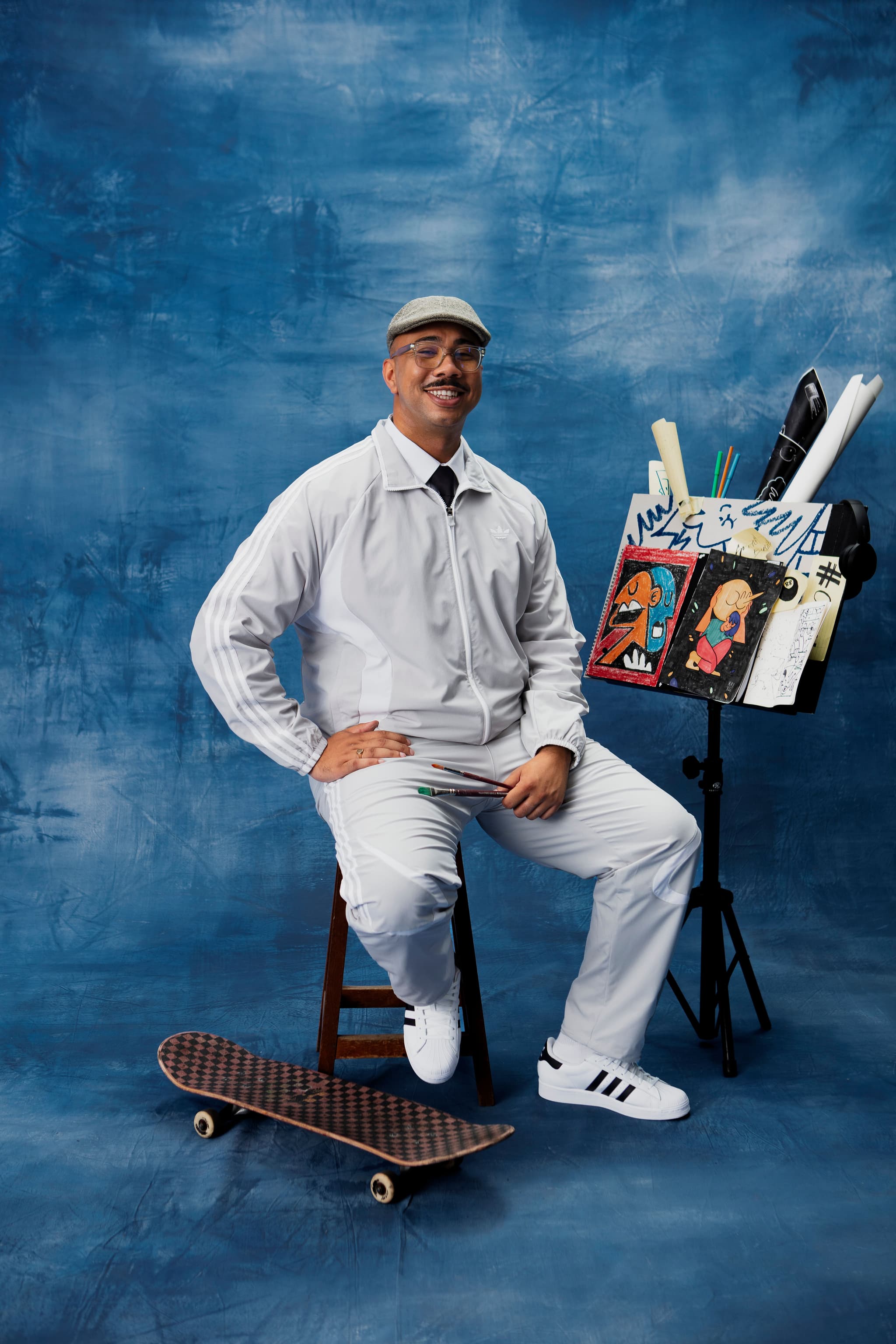 Smiling man in a white adidas tracksuit sitting with paintbrushes next to an easel with artwork and a skateboard.