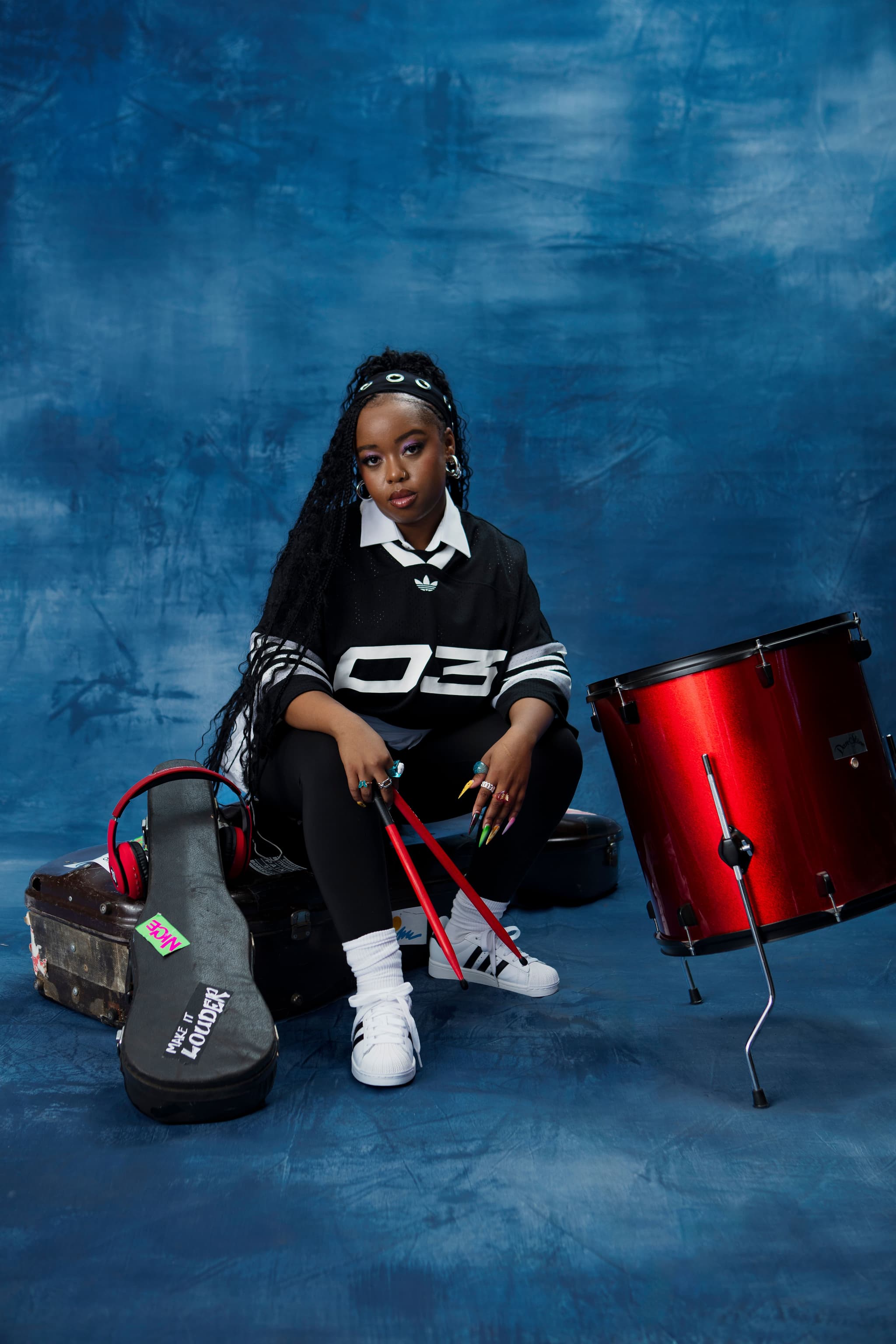 Woman in black adidas jersey holding red drumsticks, sitting on an instrument case next to a red drum on a blue background.