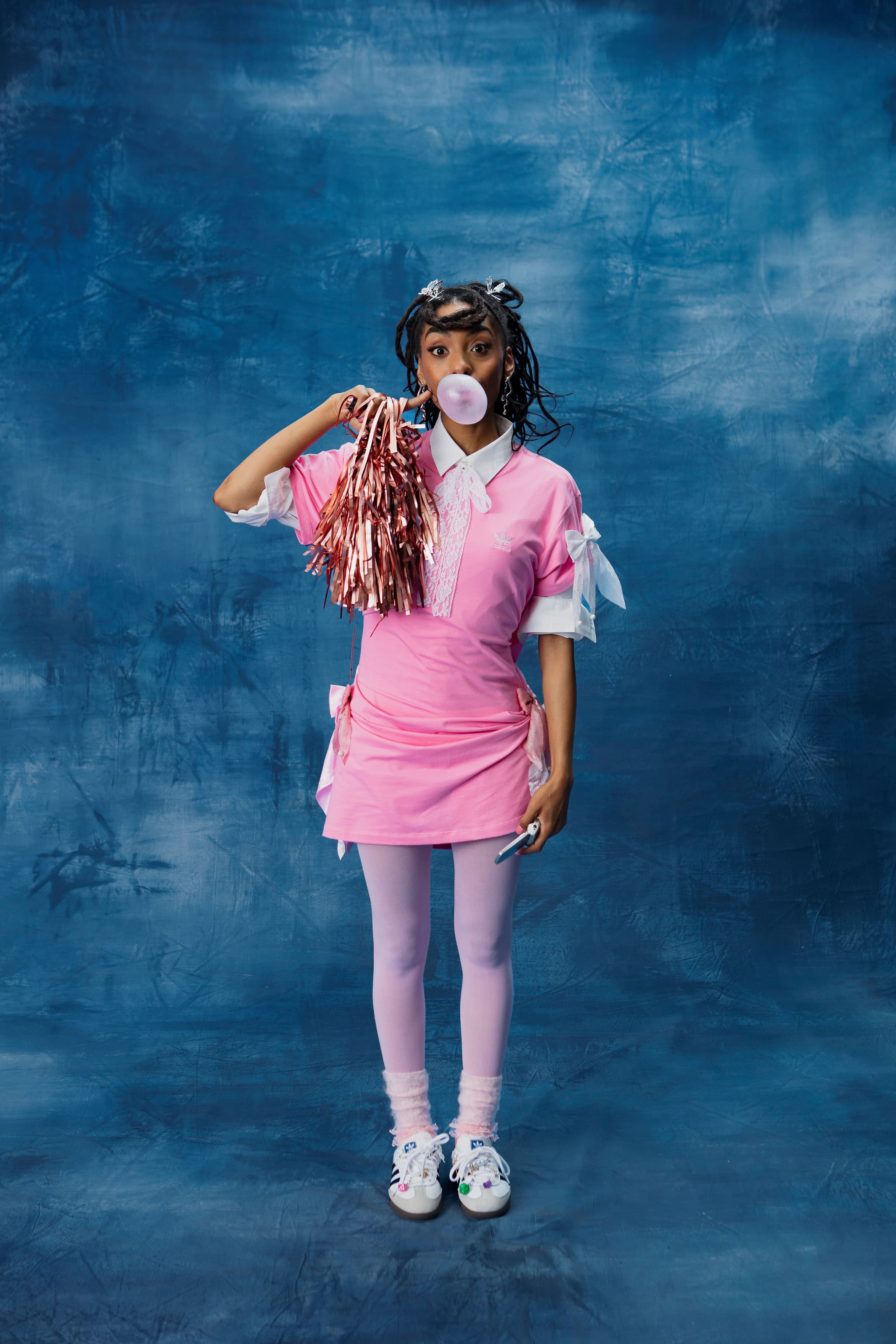 Woman in a pink dress and adidas sneakers blowing a bubblegum bubble while holding pink pom-poms on a blue background.
