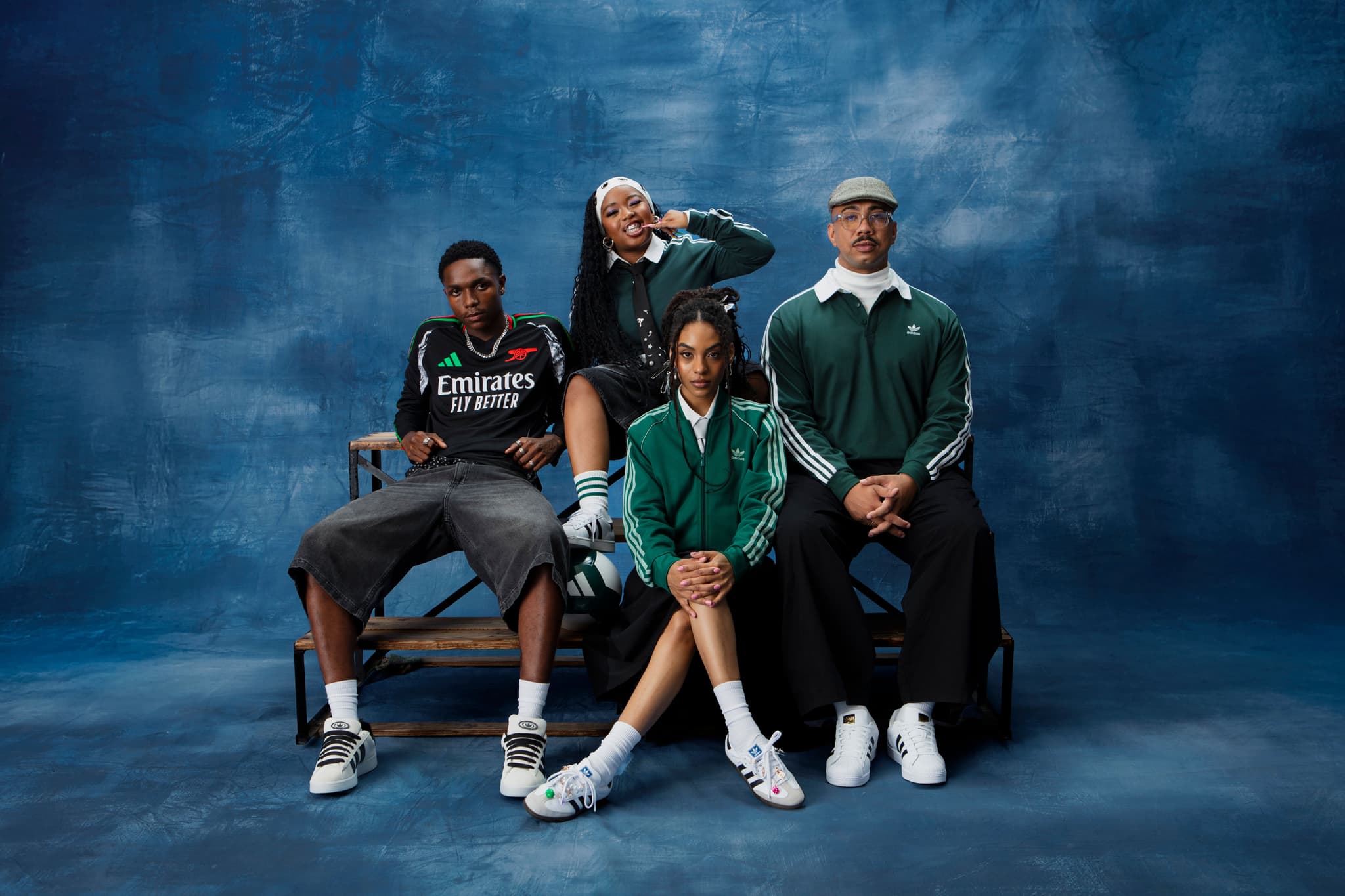Four people wearing green, black, and white adidas apparel posing together on wooden bleachers against a blue backdrop.