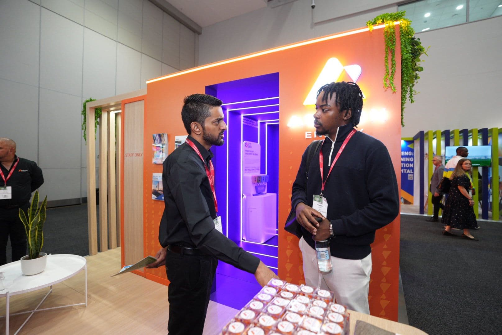 Two men engaged in conversation in front of the orange Astron Energy stand at a trade exhibition.