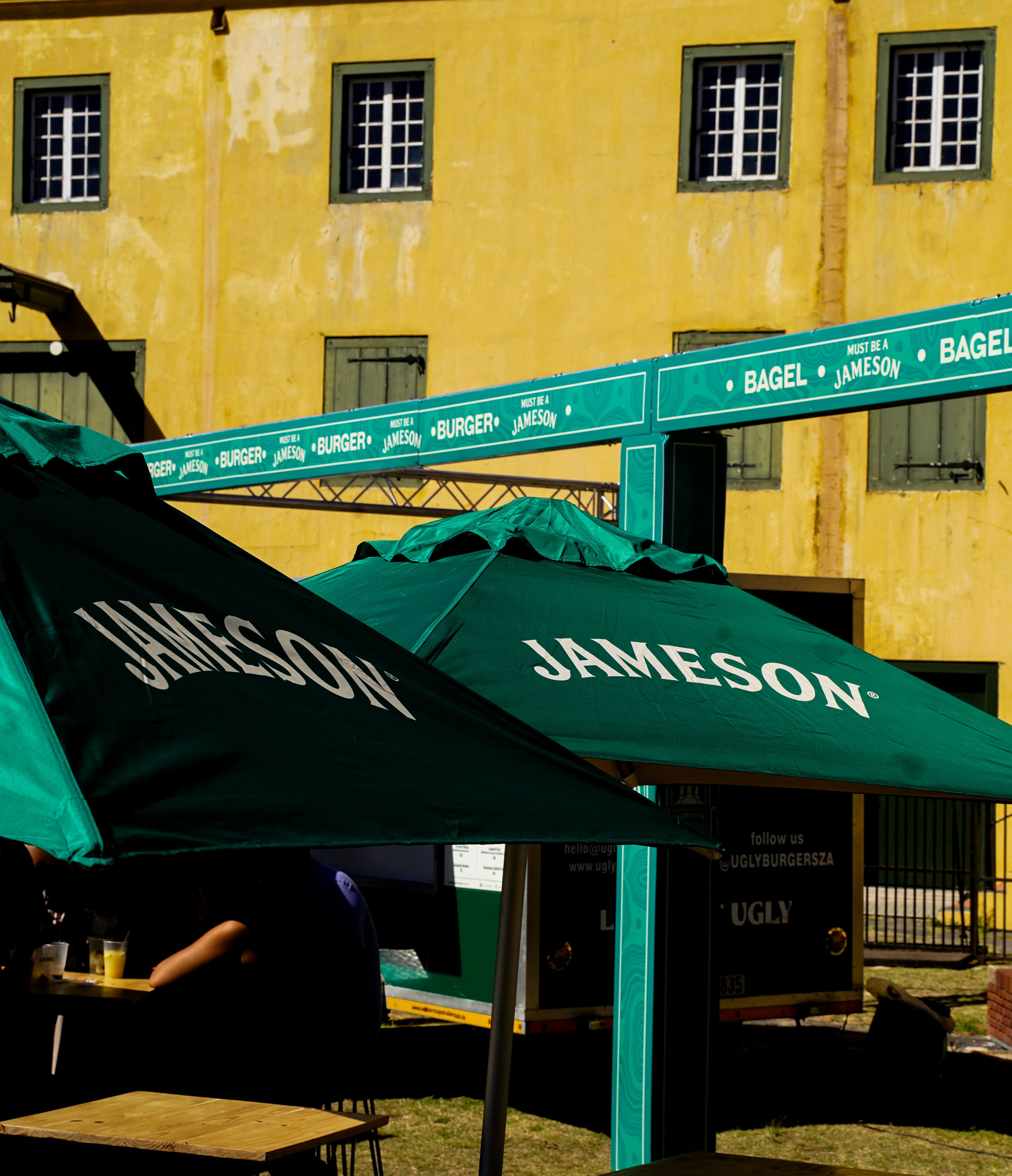 Green Jameson branded umbrellas shading an Ugly Burgers food stall at an outdoor event with a yellow building in the background.