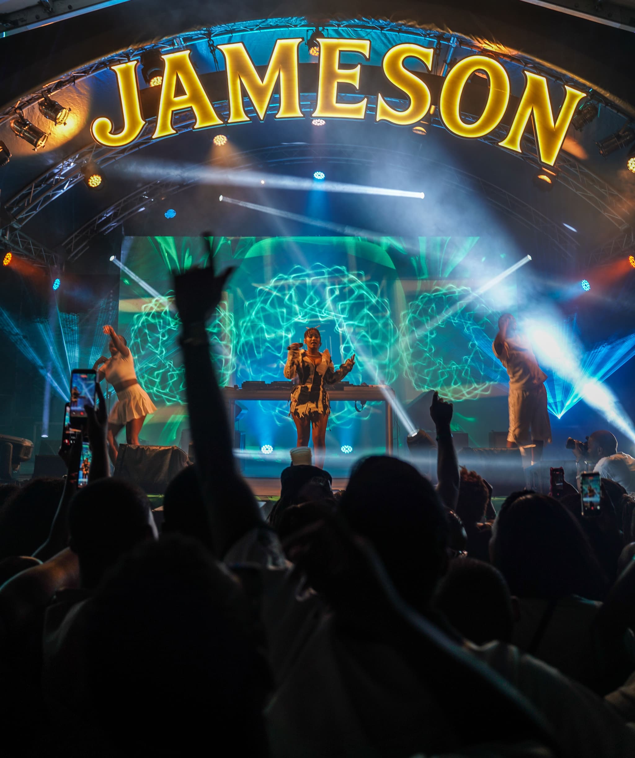 Female music artist performing on a brightly lit stage under a large Jameson arch sign to a crowd holding up phones.