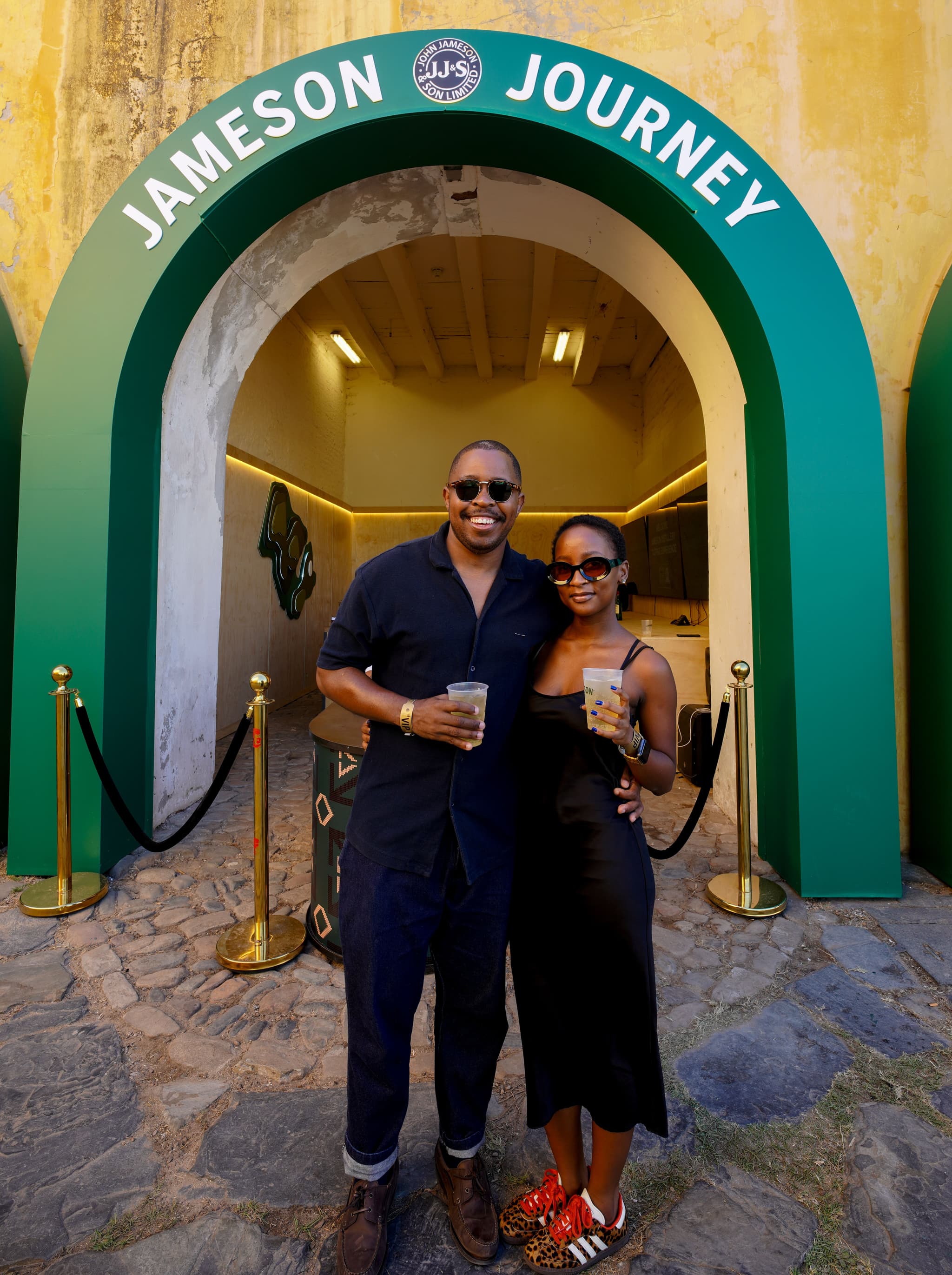 A smiling couple holding drinks poses in front of the green archway entrance to the Jameson Journey experience.