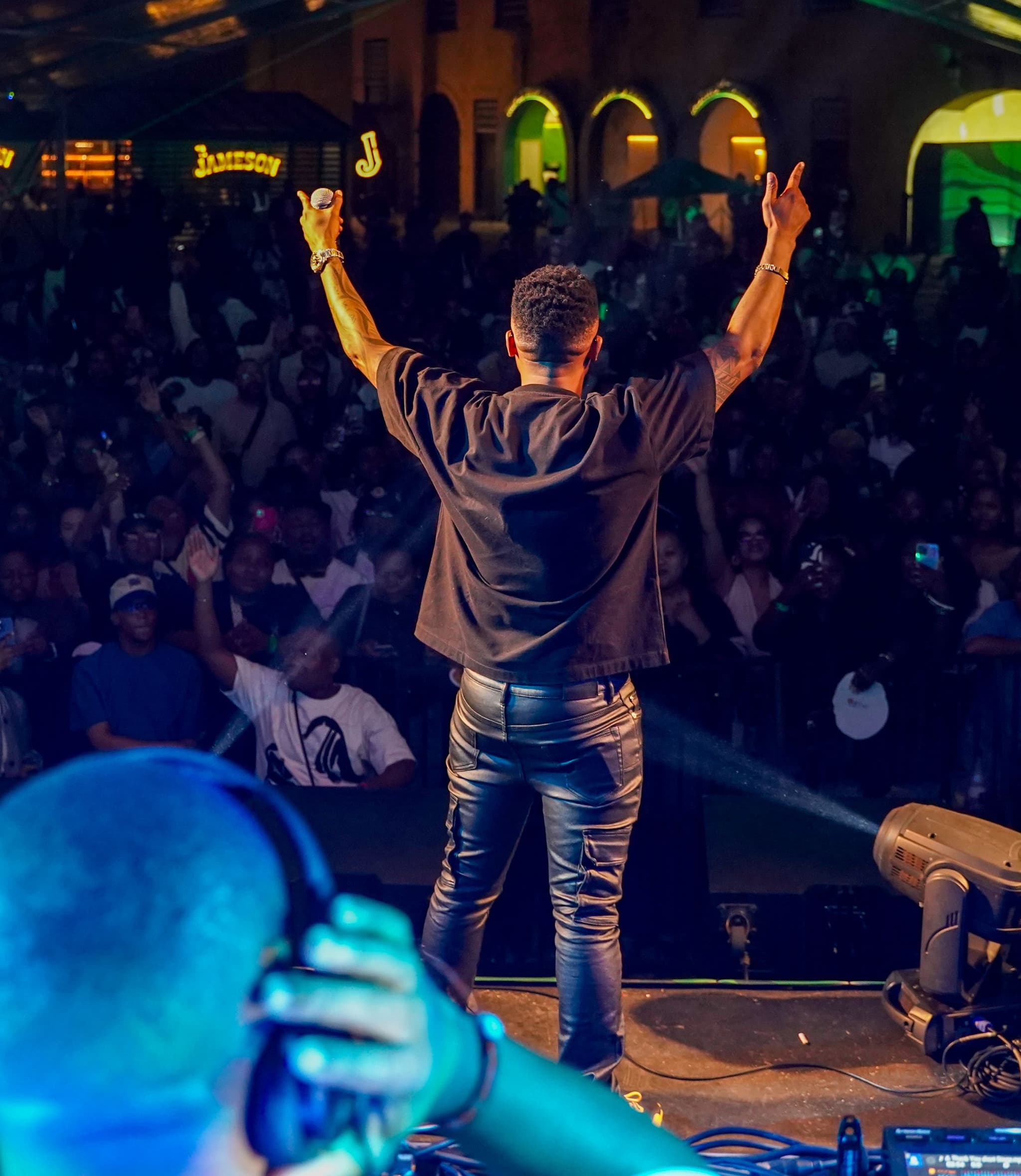 View from behind an artist on stage with arms raised, performing for a large crowd at a nighttime Jameson festival.