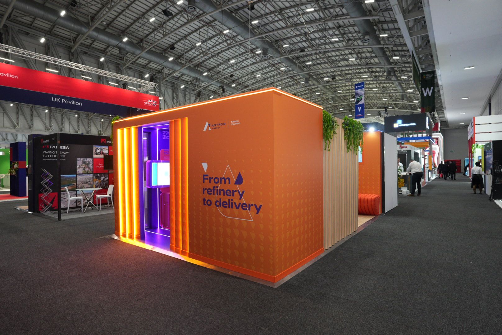 Wide view of the bright orange Astron Energy exhibition stand with "From refinery to delivery" text on the exterior wall.