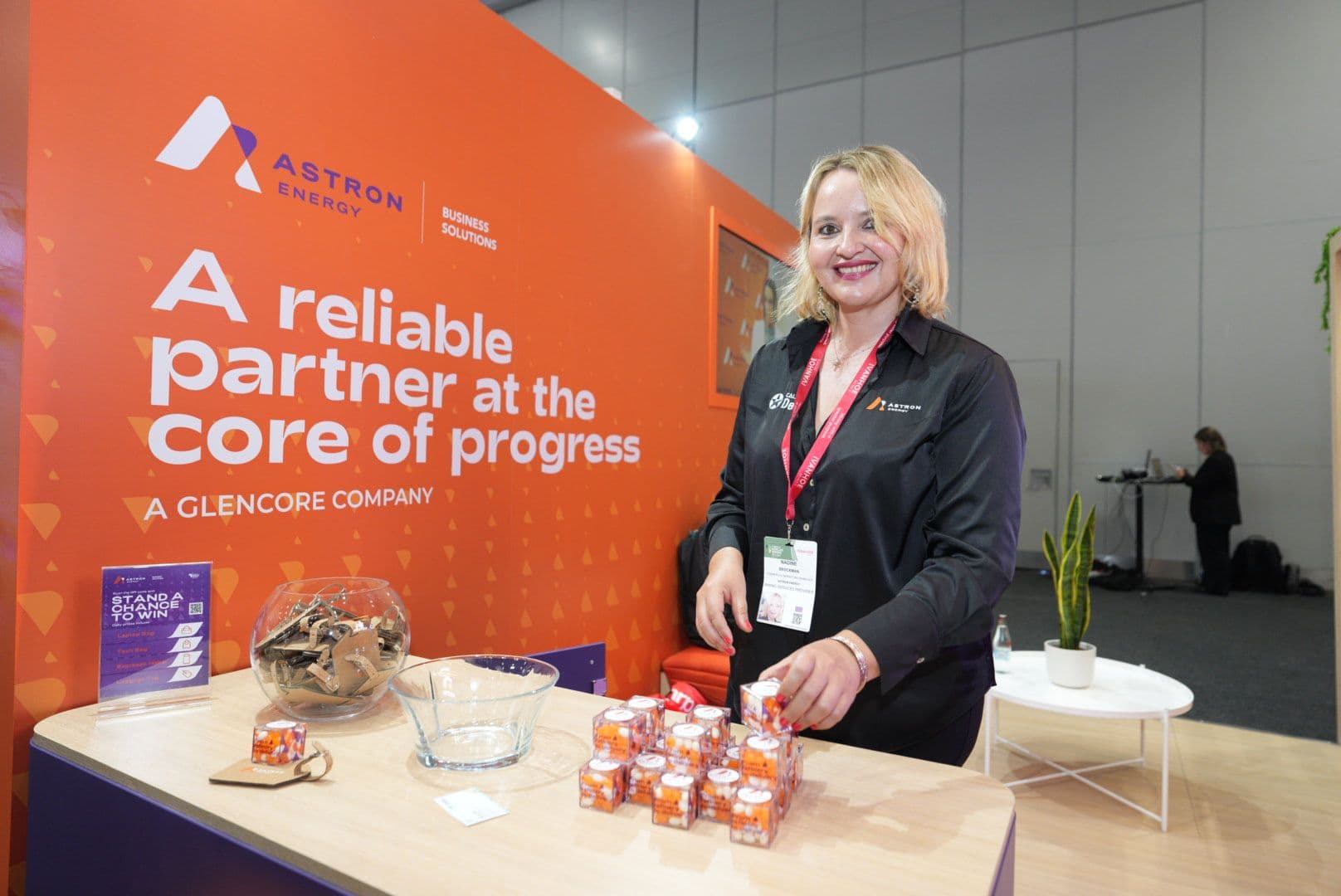 Smiling woman standing behind an Astron Energy counter displaying promotional gifts in small clear boxes.