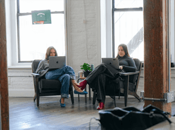 Two women working on laptops while seated in comfortable armchairs at the IMA US office.