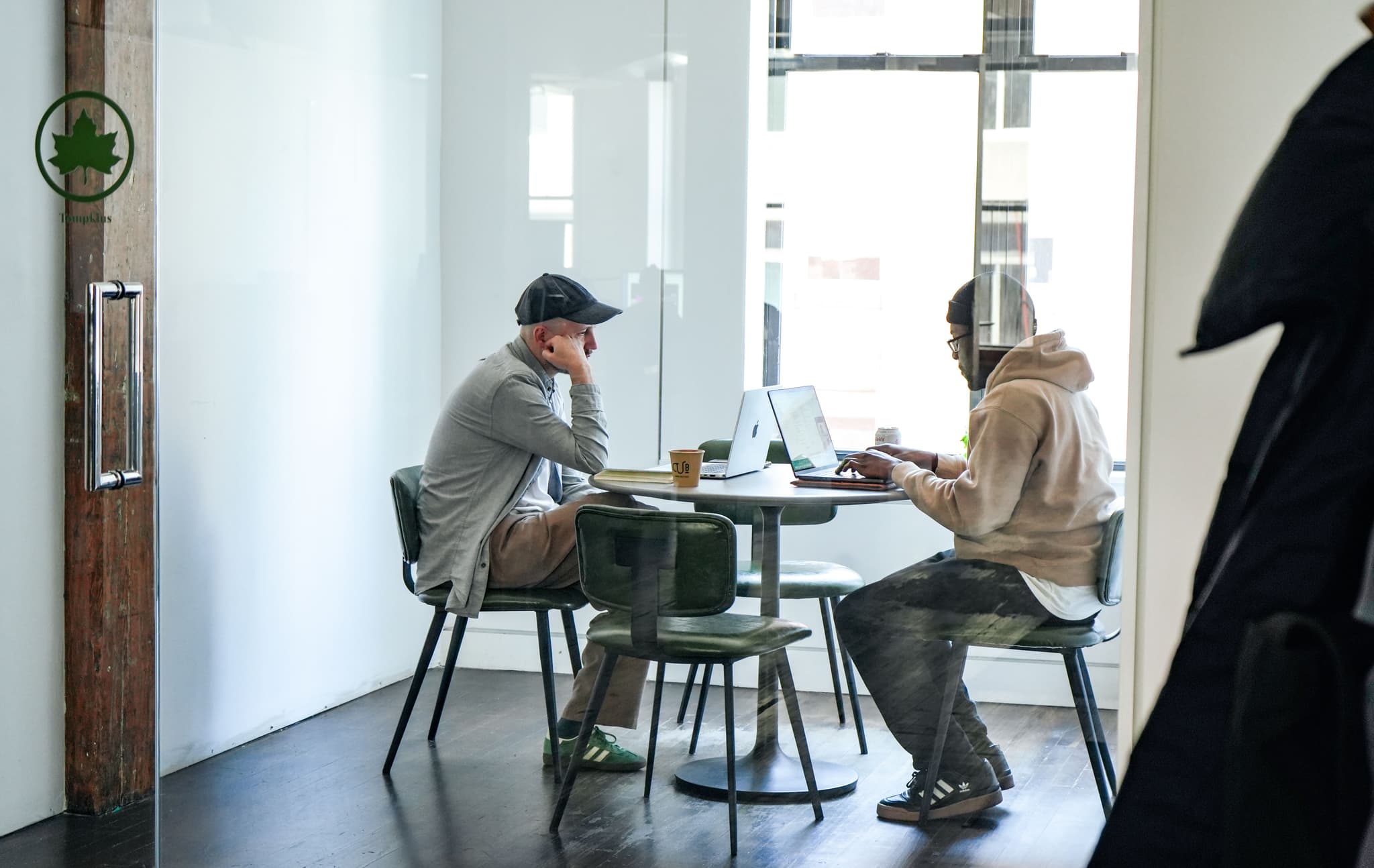 Two men working on laptops at a round table inside a glass meeting room at the IMA New York office.