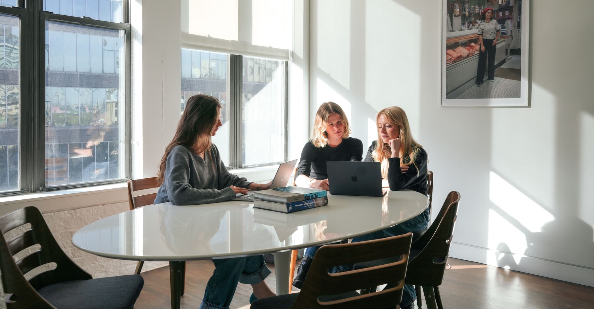 Three female employees working together around a laptop at a sunlit table in the IMA New York office.