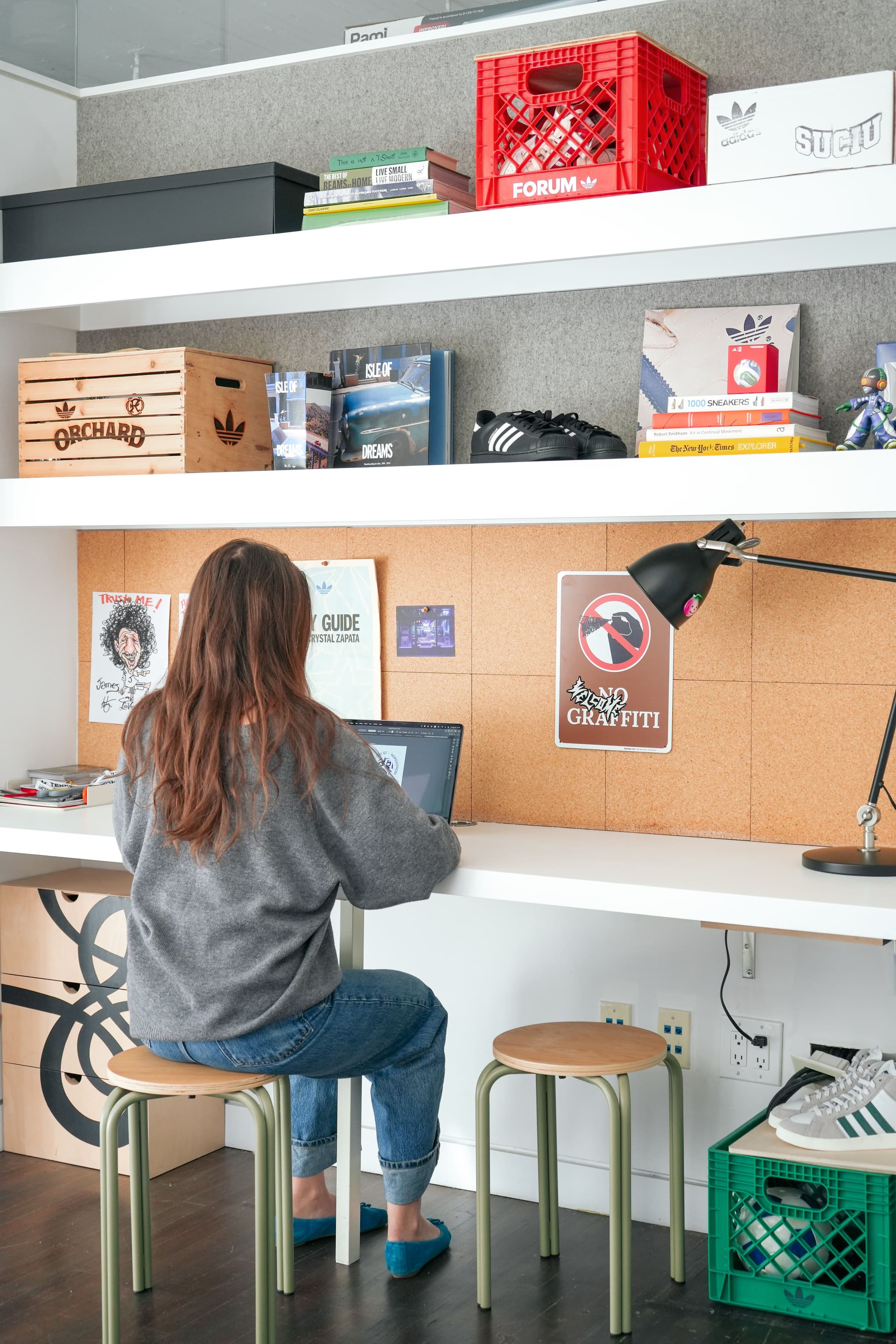 Woman working on a laptop at a desk with a corkboard and decorated shelves in the IMA New York office workspace.