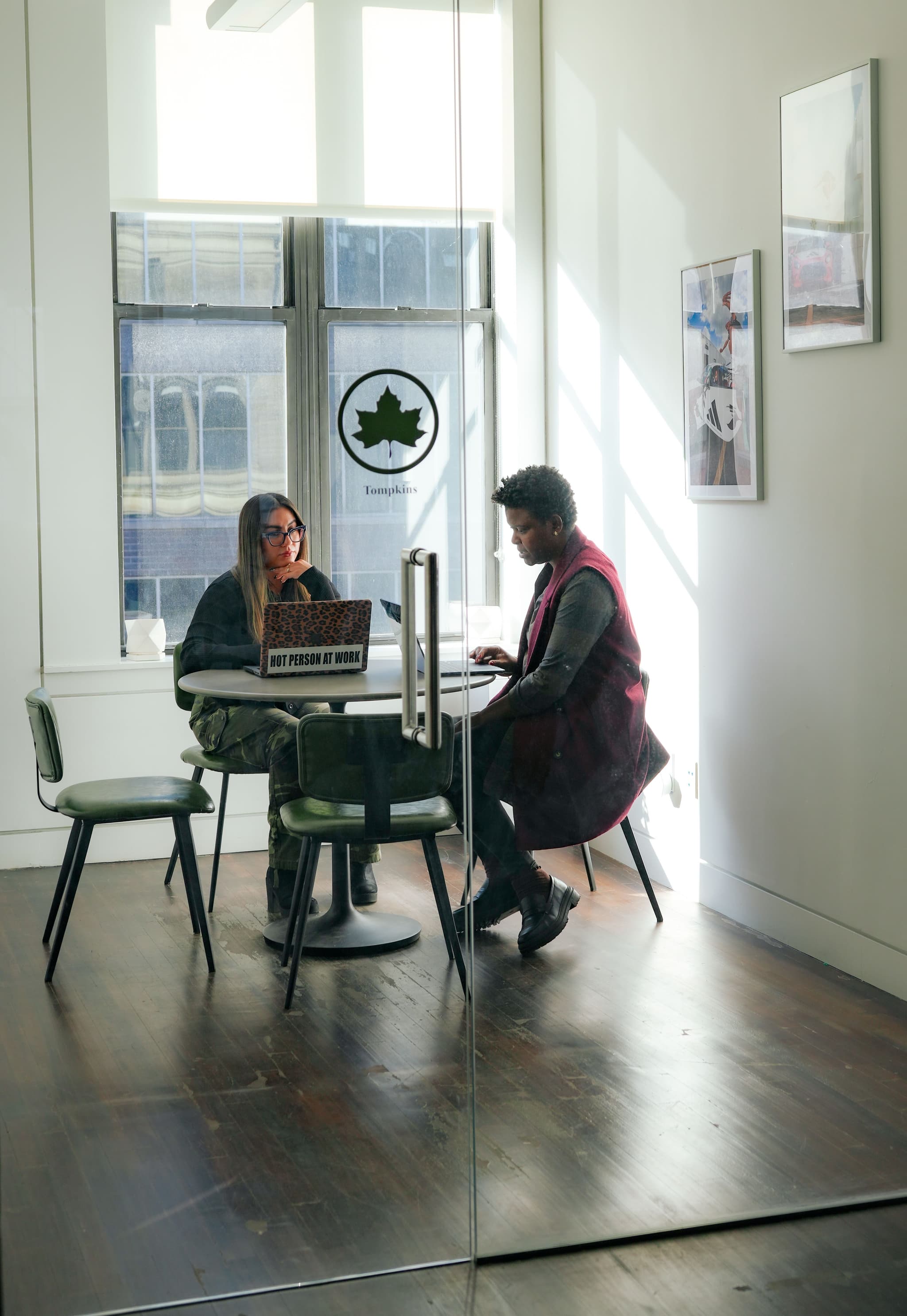 Two women working on laptops at a small round table inside the glass Tompkins meeting room at the IMA New York office.
