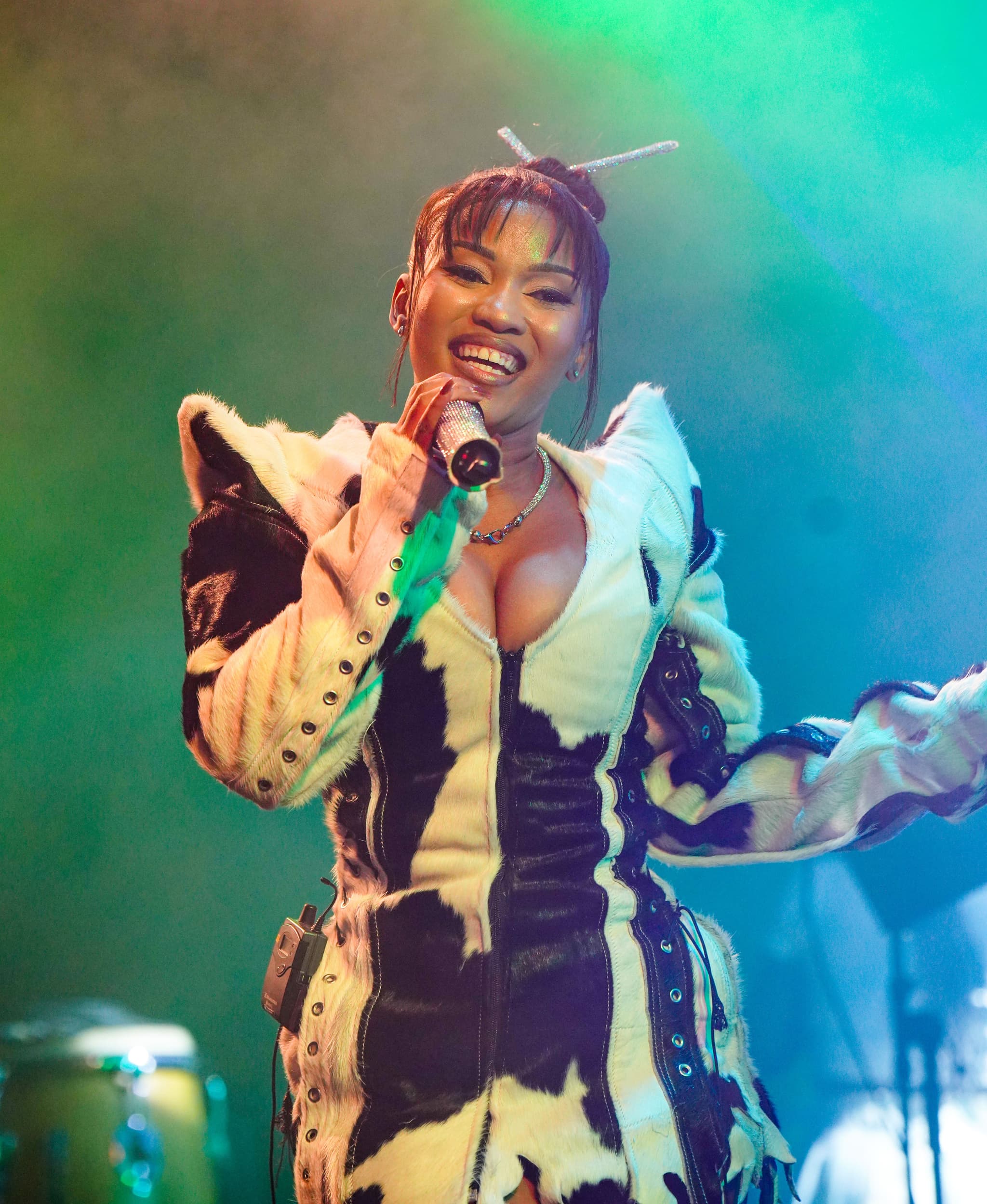 Female artist smiling and singing into a microphone on stage, wearing a striking black and white cow-print outfit.