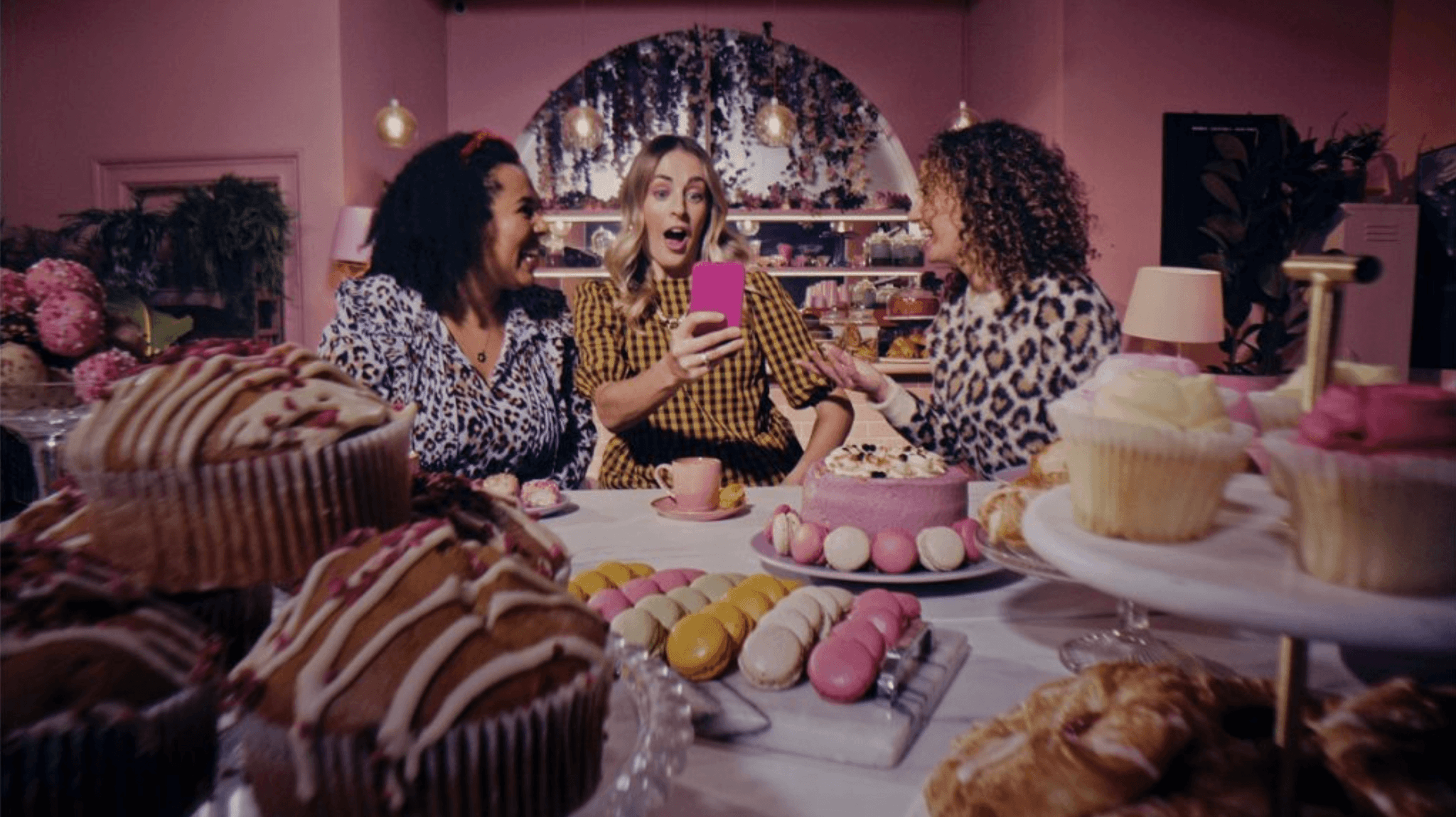 Three women in a pink cafe look excited and surprised at a smartphone, sitting at a table filled with cakes and pastries.