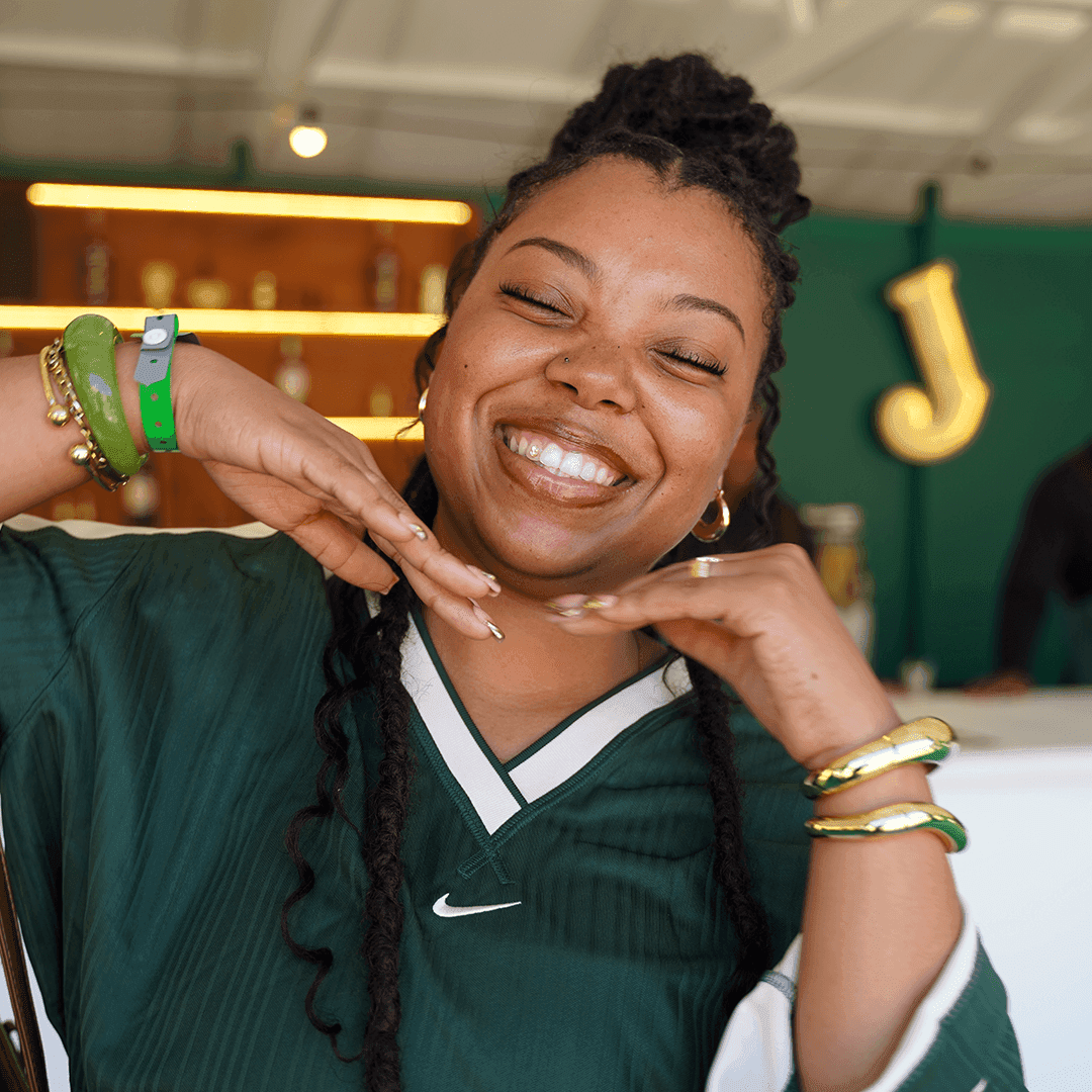 A joyful woman in a green Nike jersey and gold bangles smiling with her hands framing her face at a Jameson event.