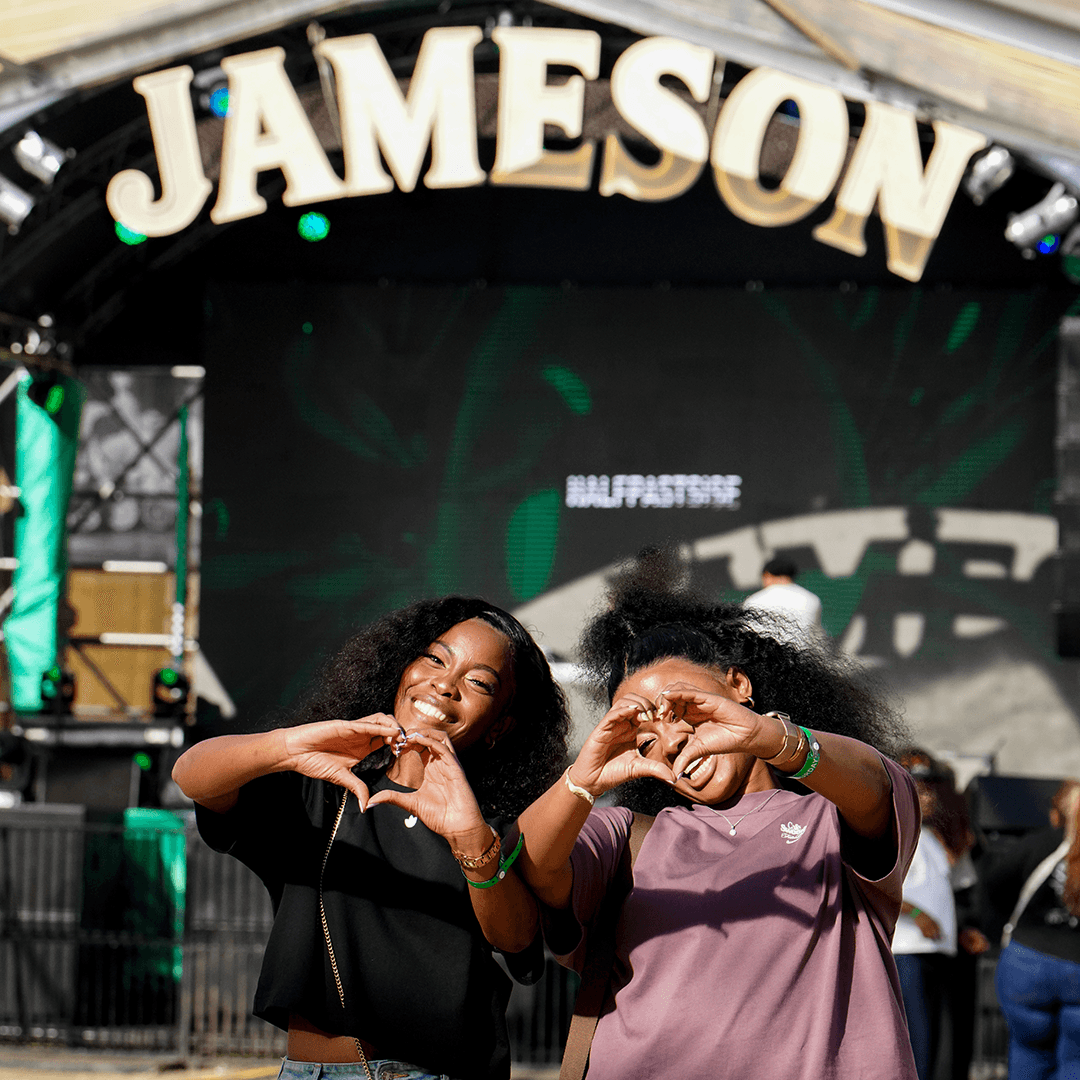 Two smiling women making heart shapes with their hands in front of the main Jameson festival stage during the day.
