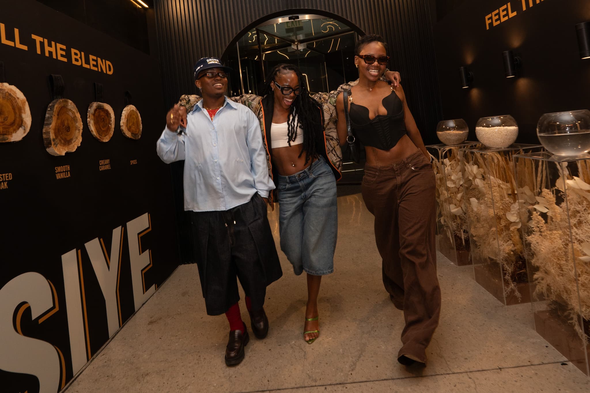 Three smiling friends walking arm-in-arm through a branded event corridor with "Feel the Blend" text.