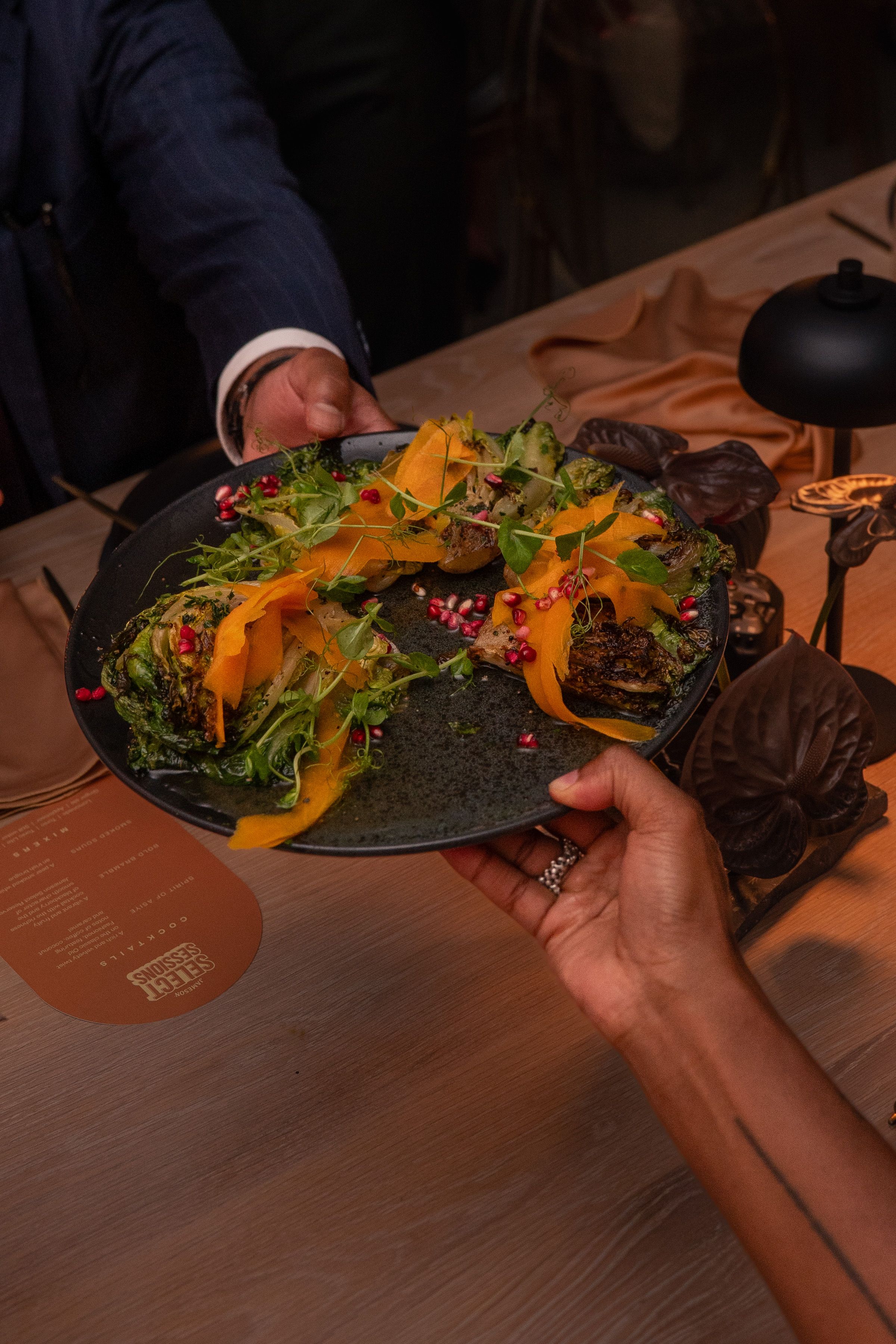 Hand holding a dark plate with a gourmet salad topped with pomegranate seeds at a Select Sessions event.