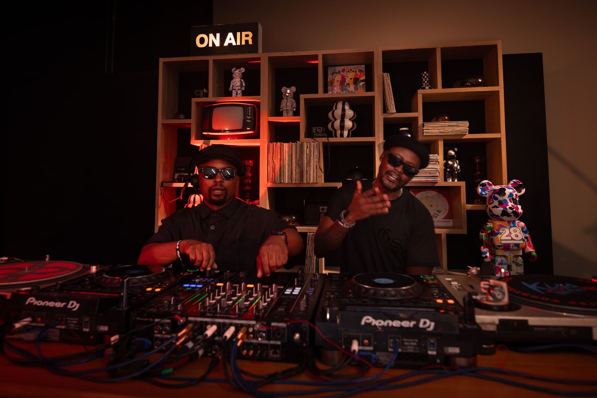 Two DJs in sunglasses performing on Pioneer turntables in a studio setting with an ON AIR sign.
