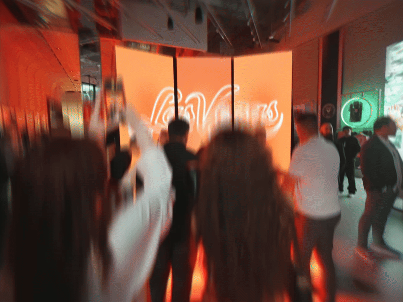 Blurred crowd in front of a glowing Las Vegas neon sign at a dimly lit event.