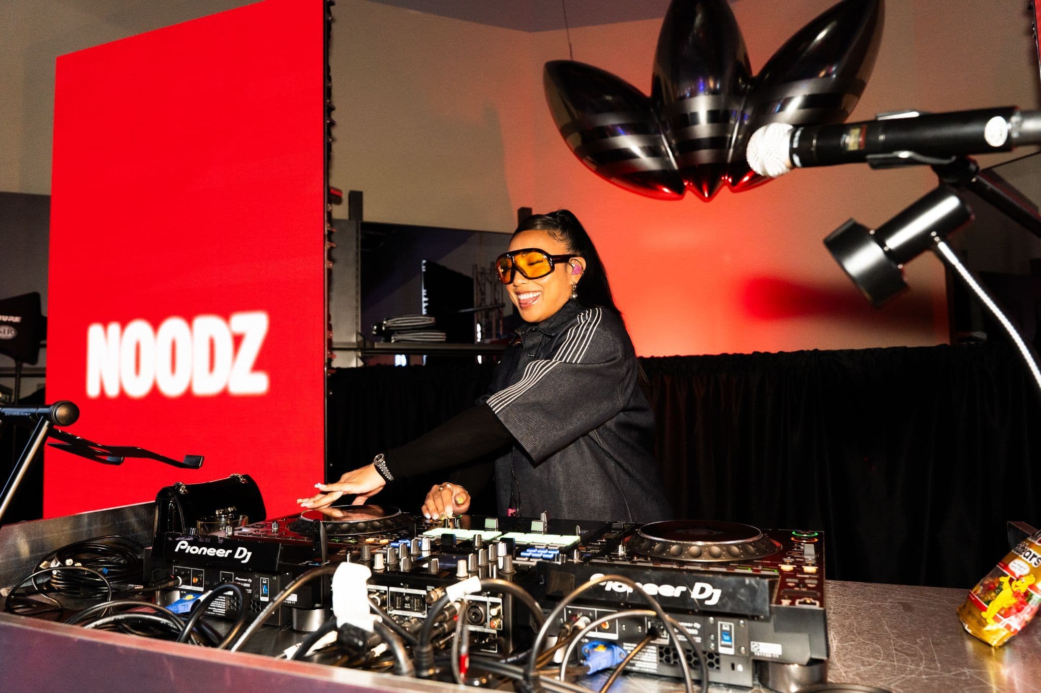 A DJ wearing sunglasses performing behind a mixing console at a red-lit venue.