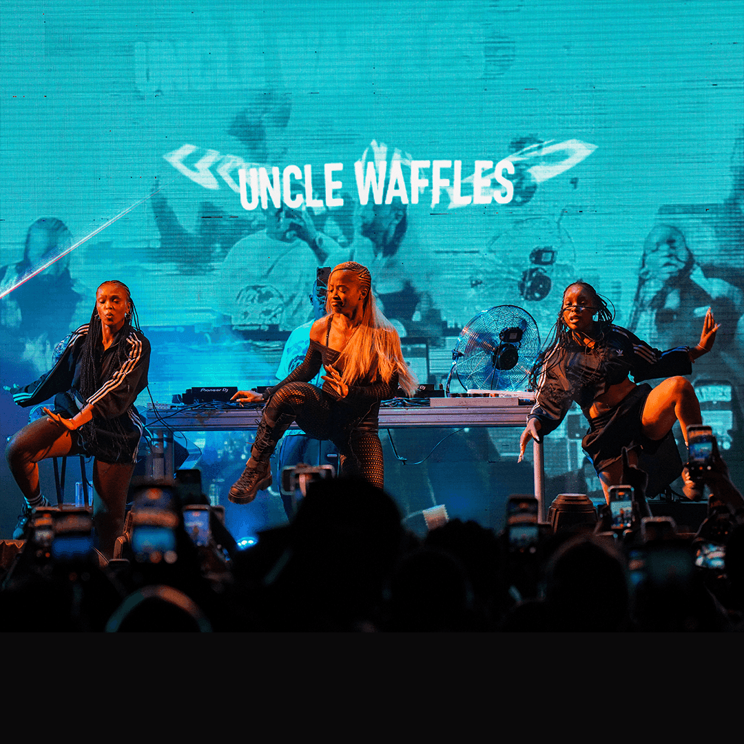 Uncle Waffles and two dancers performing energetically on stage in front of a large crowd holding up smartphones.