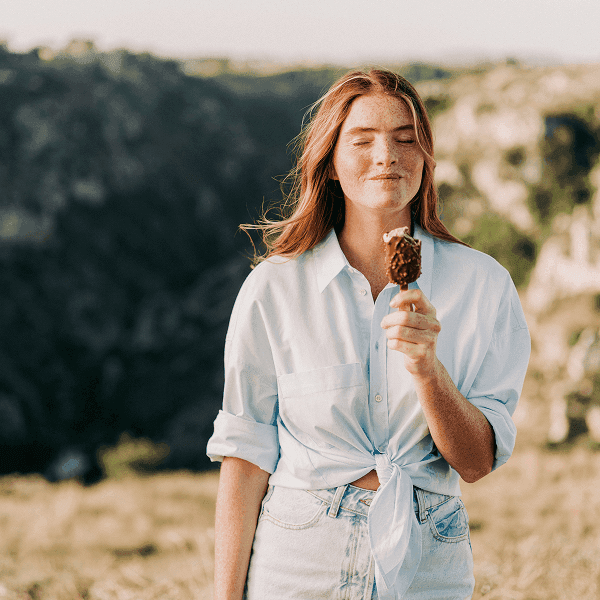 A woman in a light blue button-down shirt holding a chocolate ice cream cone in a natural setting.