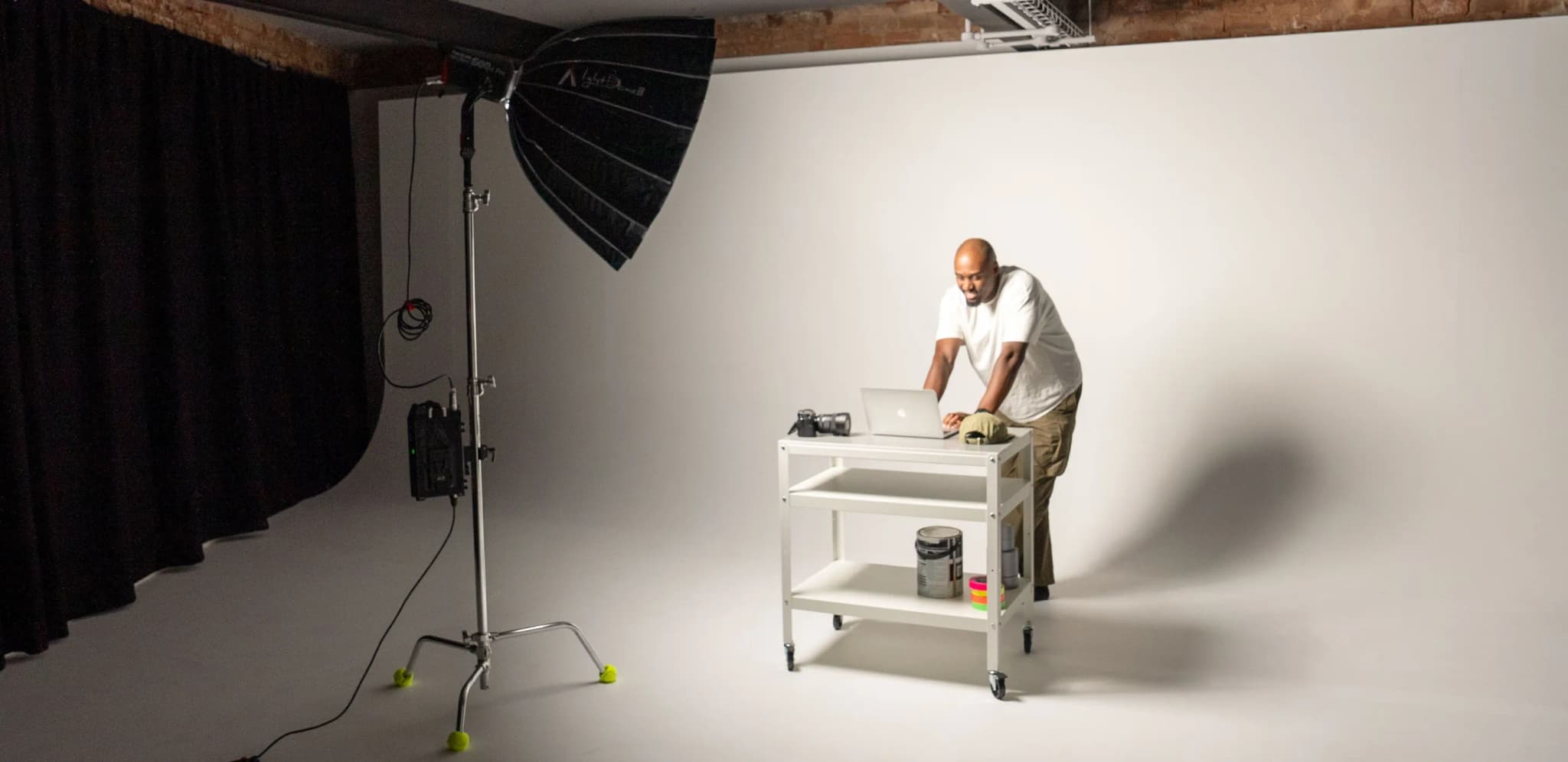 A photographer using a laptop on a white rolling cart in a studio with a large softbox light.