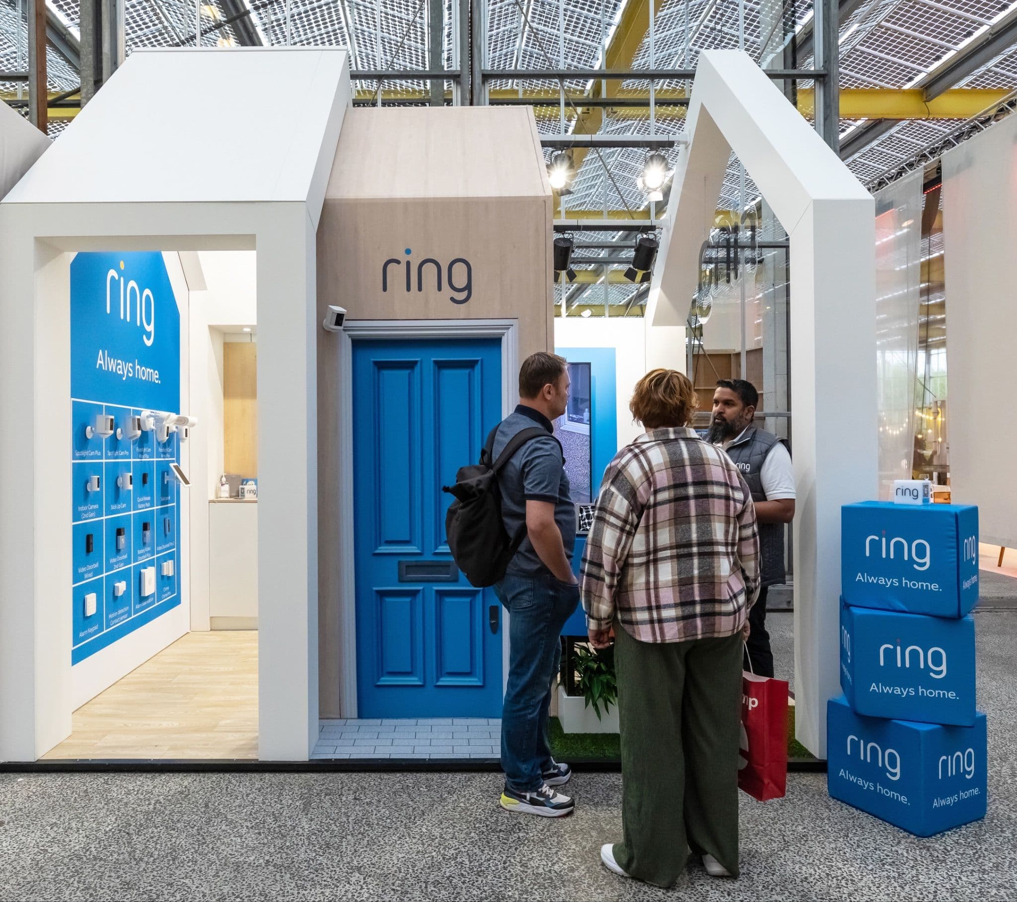 Visitors chatting at a house-shaped Ring exhibition booth featuring a blue door and stacked branded boxes.