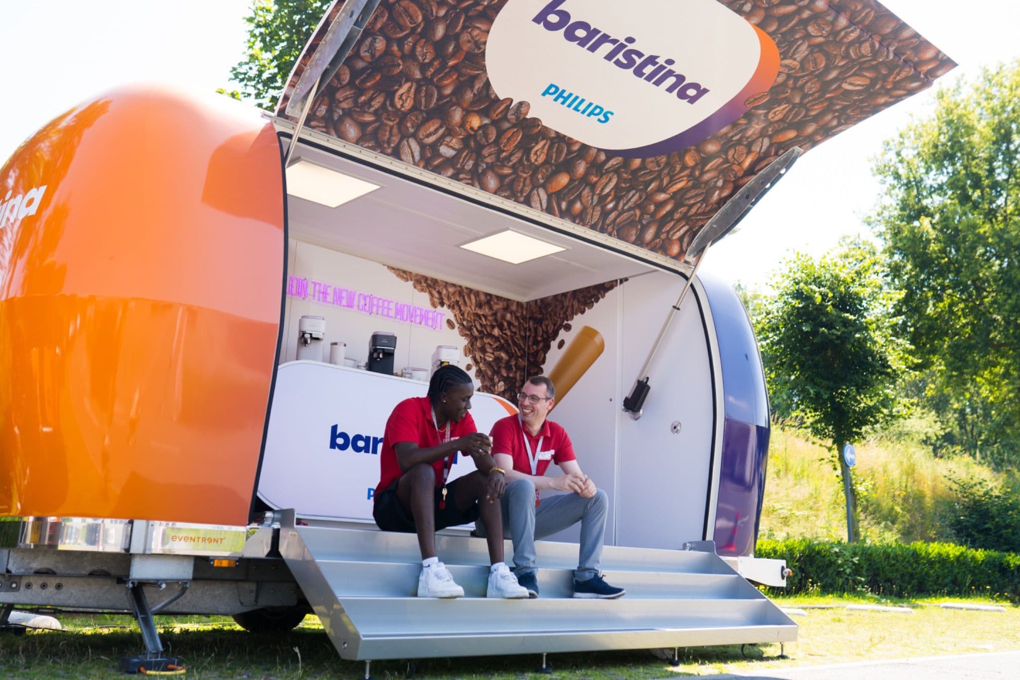 Two staff members in red shirts sitting and chatting on the steps of an open Philips Baristina mobile coffee trailer.