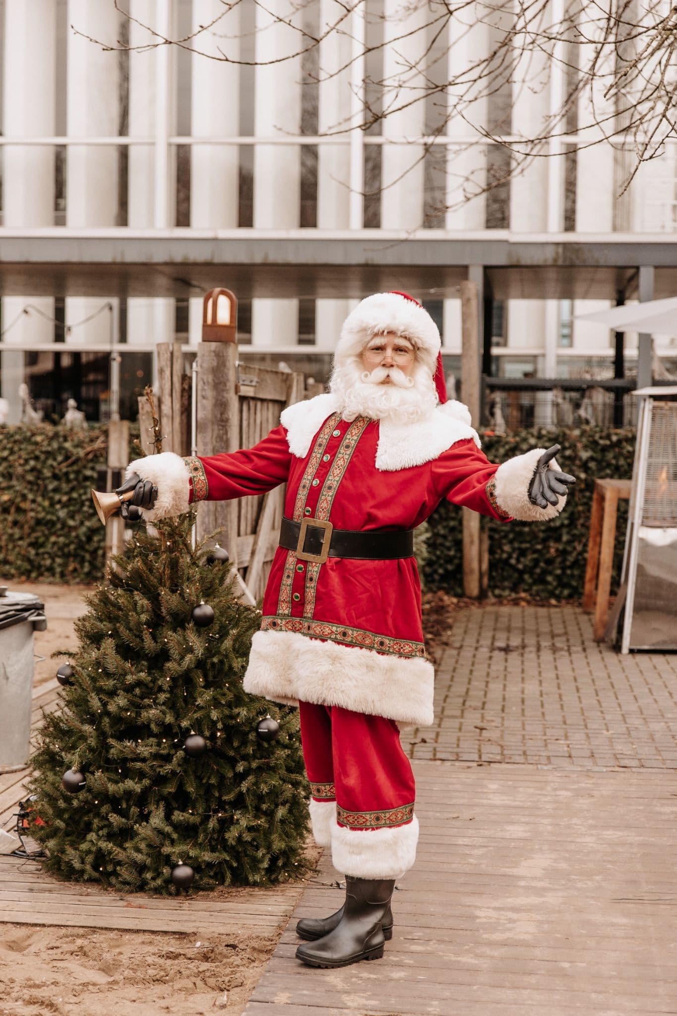 Santa Claus holding a bell standing next to a Christmas tree decorated with black baubles.