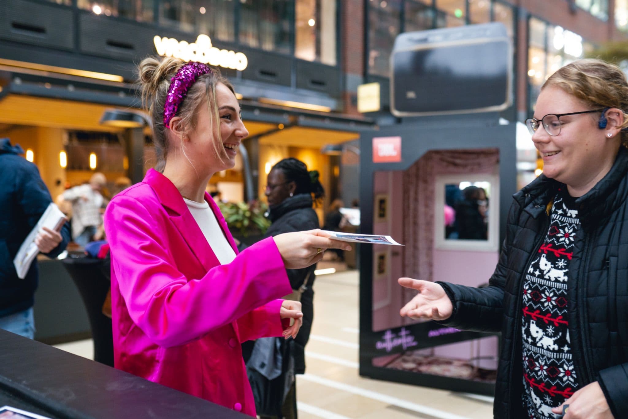 Smiling promotional staff member in a pink blazer handing a photo print to a visitor at a JBL mall activation event.