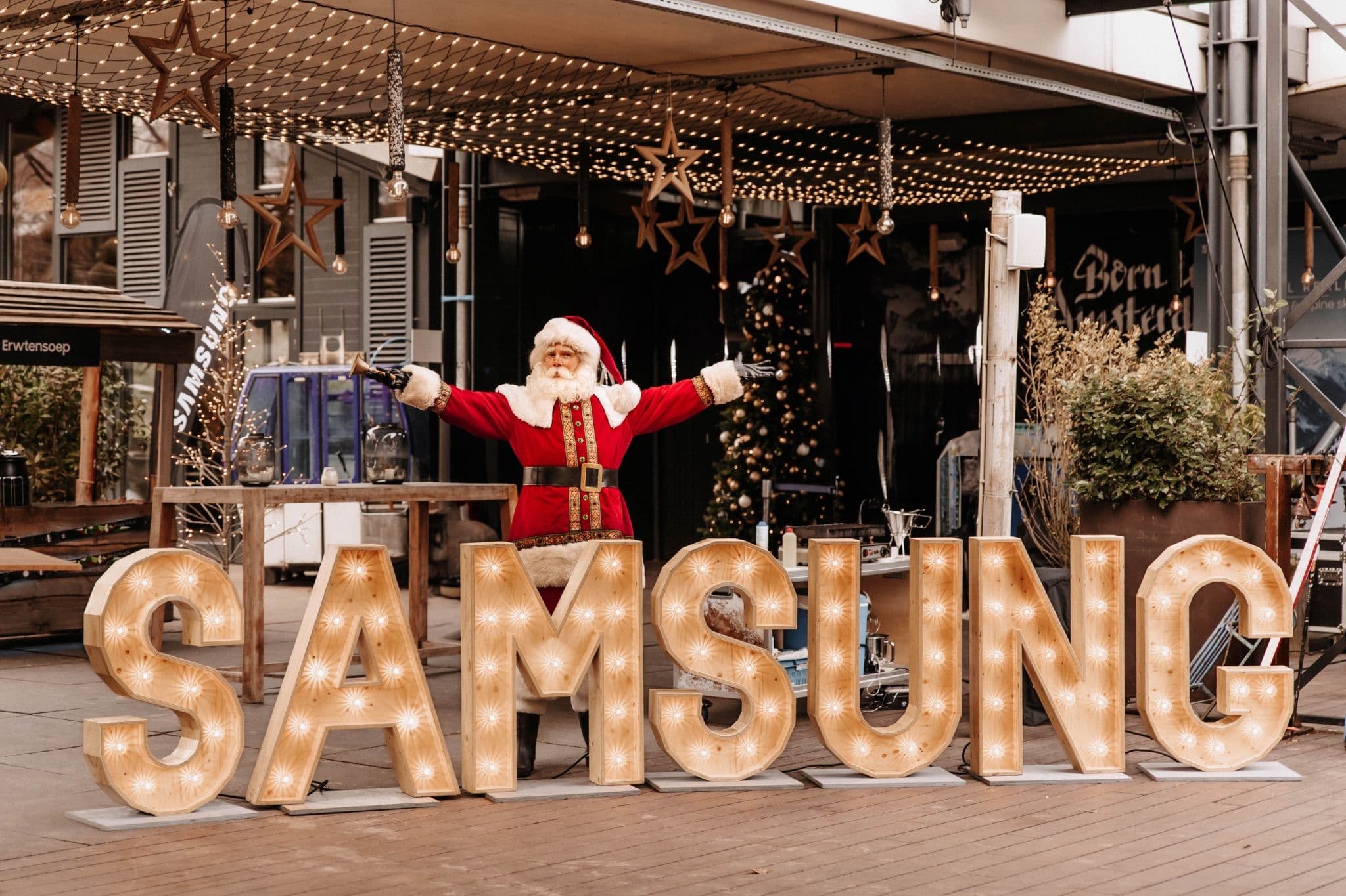 Santa Claus celebrating behind large illuminated wooden letters spelling SAMSUNG at an outdoor venue.
