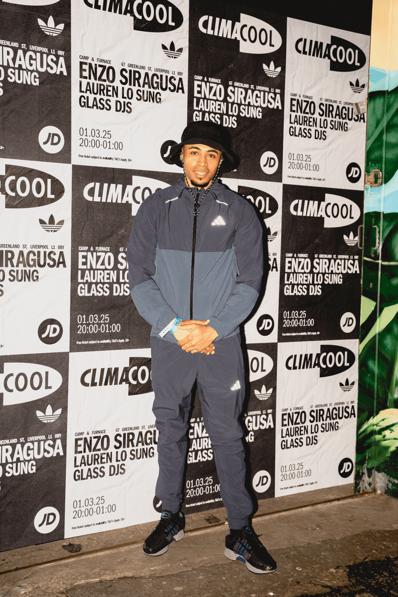 Man in an Adidas tracksuit posing before a Climacool event poster wall featuring Enzo Siragusa and JD branding.