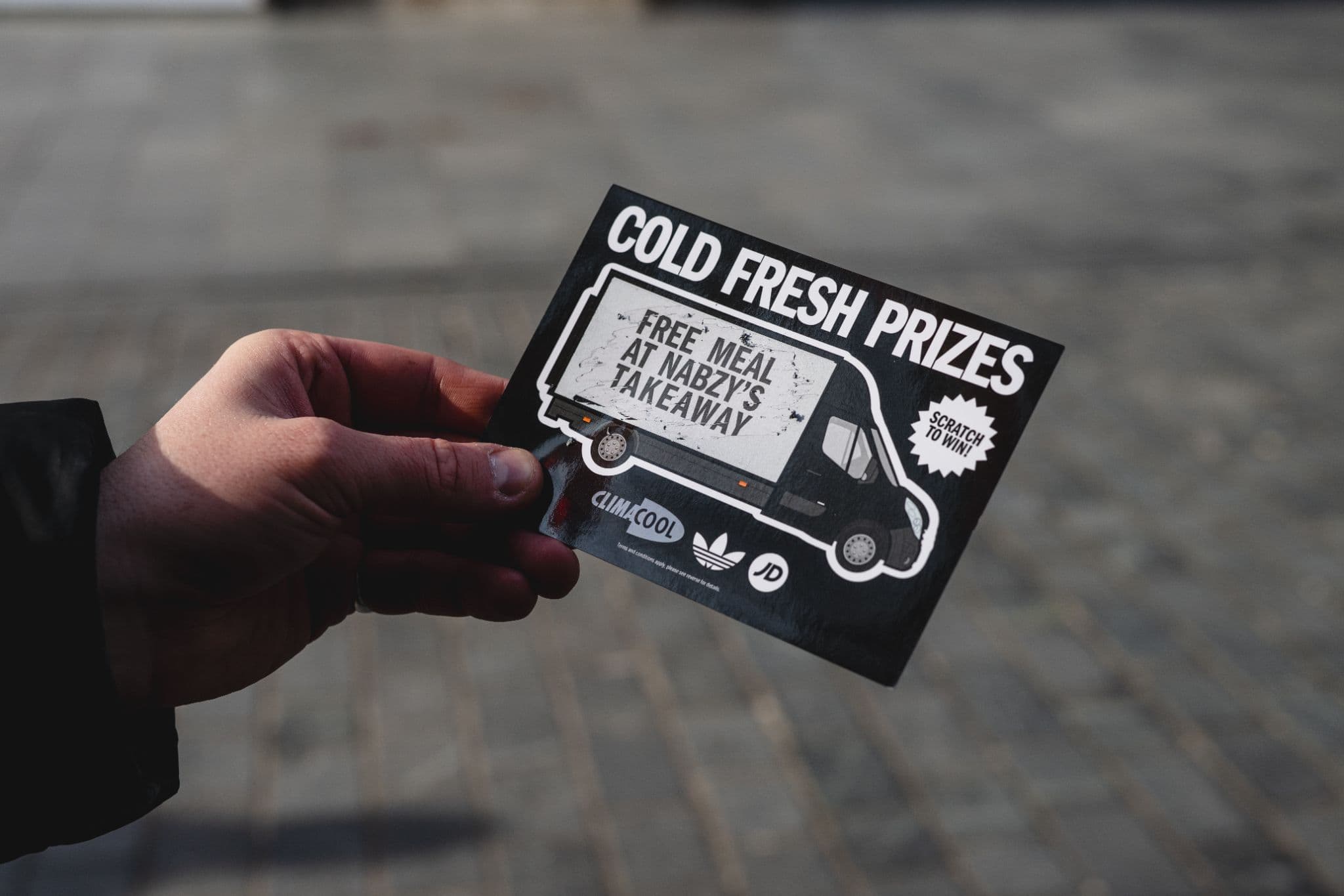 Hand holding a "Cold Fresh Prizes" scratch card offering a free meal at Nabzy's, featuring Adidas and JD logos.