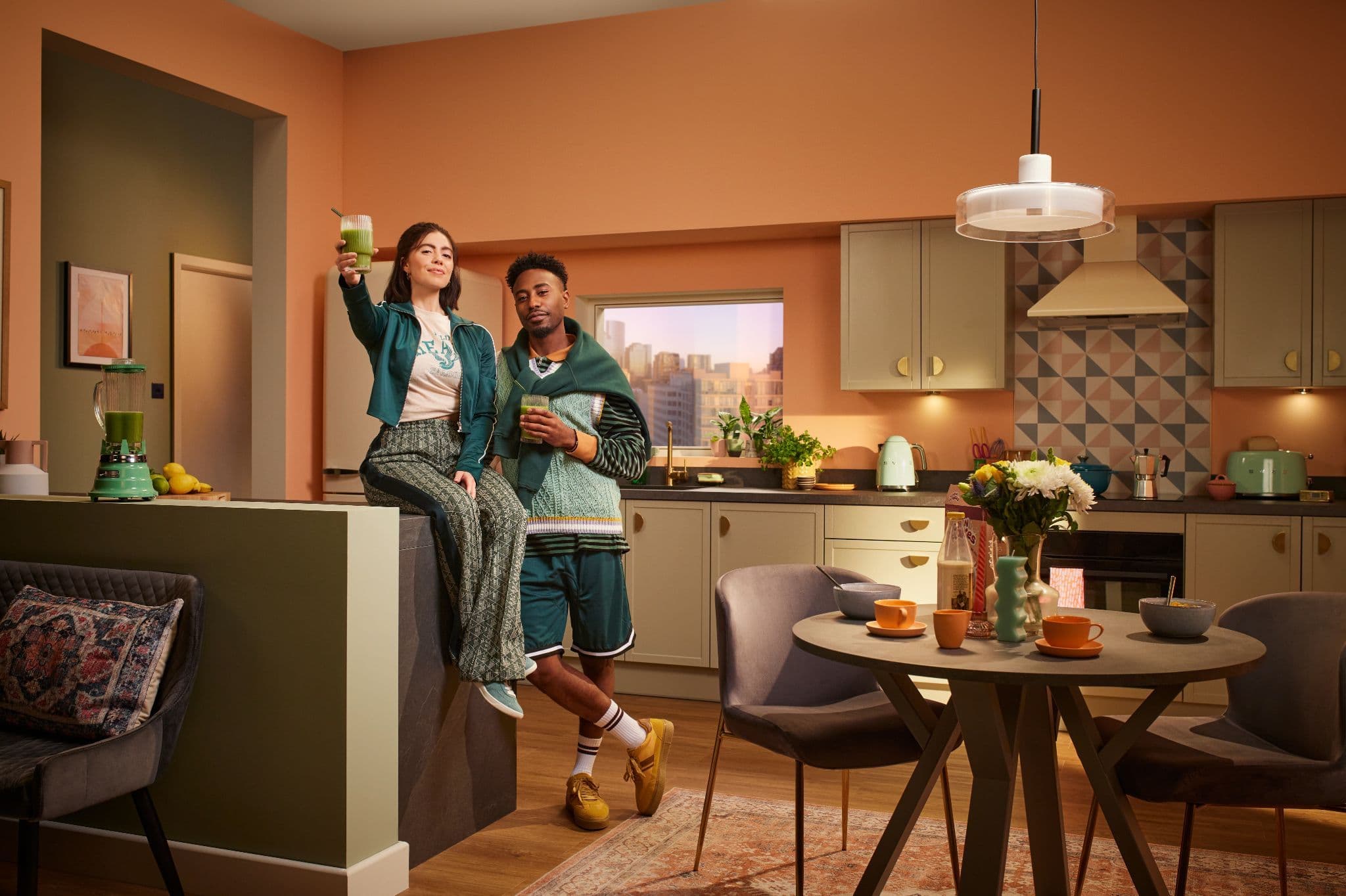 Stylish couple holding green smoothies posing in a modern kitchen with sage green cabinets and pink walls.